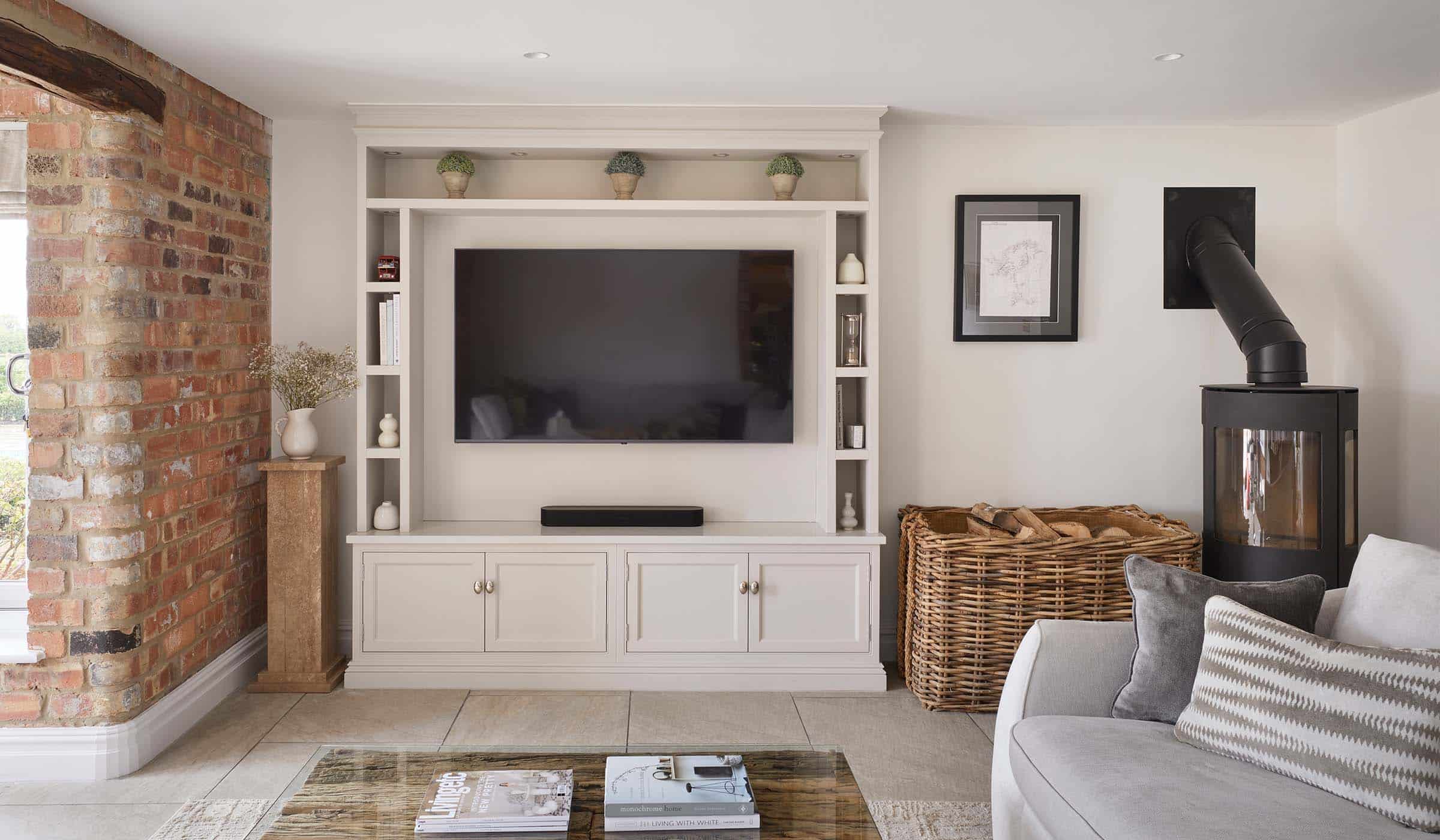 A bespoke media wall in French Grey. The bespoke floor-to-ceiling media wall has Edwardian-styled profiling that stands parallel to the exposed brick wall on its left. The mounted flat screen television stands in the middle of cabinetry crafted with left and right side-shelving including a mantel along the top with three ceramic plants spaced equally along it. Underneath, cupboards with Armac Martin handles sit flush to the cabinetry. A wicker log basket stands to the right of the media wall along with a black log burner, while a tall plinth with ceramic jug on top stands between the media wall and the exposed brick to the left.