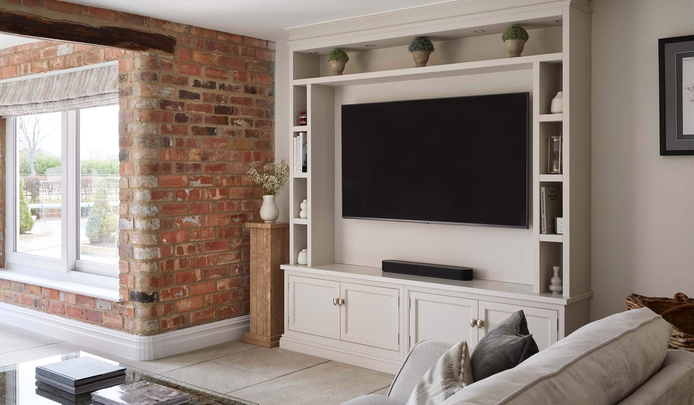 A bespoke media wall in French Grey. The bespoke floor-to-ceiling media wall has Edwardian-styled profiling that stands parallel to the exposed brick wall on its left. The mounted flat screen television stands in the middle of cabinetry crafted with left and right side-shelving including a mantel along the top with three ceramic plants spaced equally along it. Underneath, cupboards with Armac Martin handles sit flush to the cabinetry. A wicker log basket stands to the right of the media wall while a tall plinth with ceramic jug on top stands between the media wall and the exposed brick to the left. Around the corner of the exposed brick, the tall window lets in a great deal of light from the manicured view beyond.