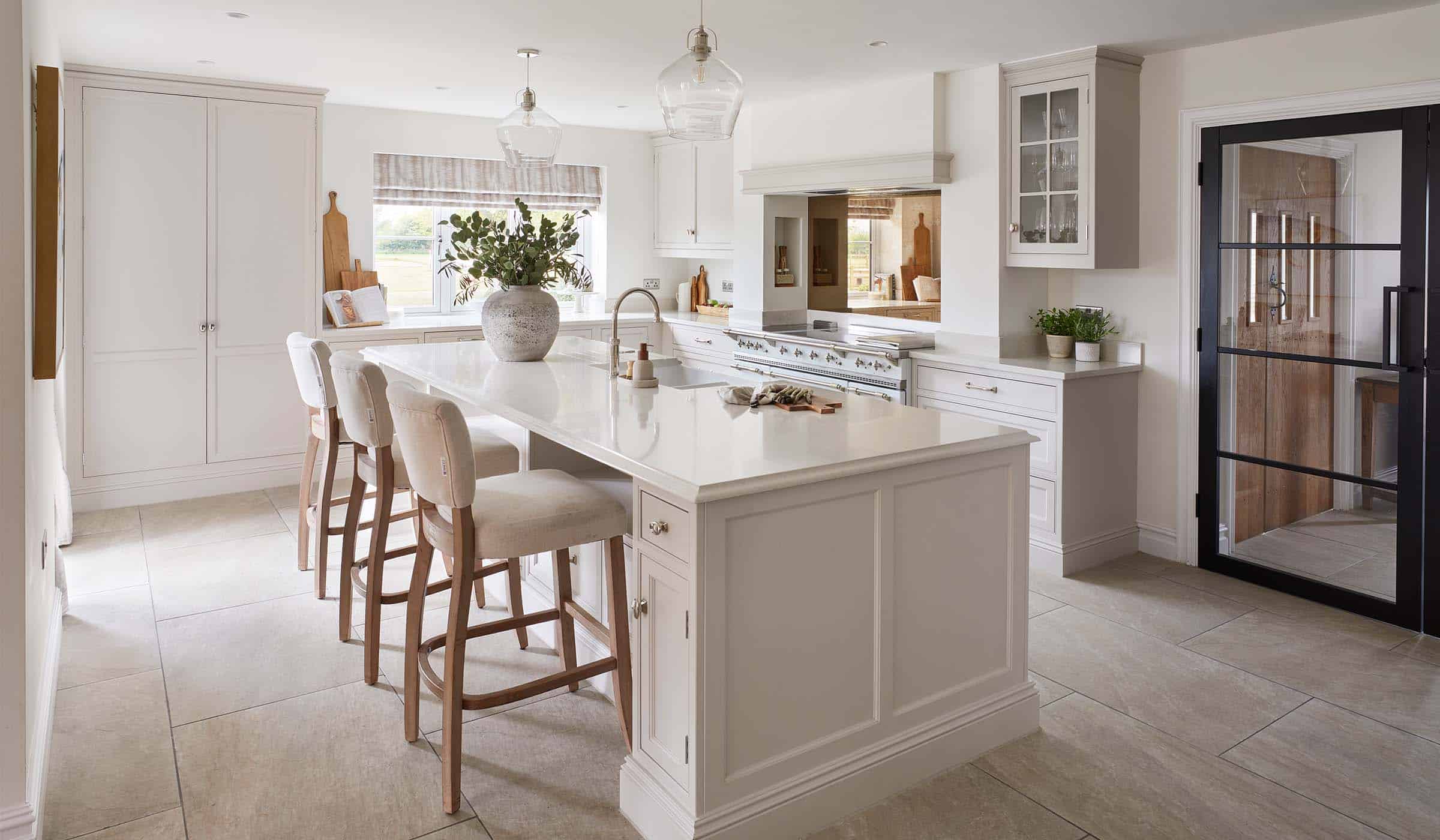 The Fox Traditional Shaker Kitchen Project A traditional shaker kitchen in cream. A floor-to-island cabinet oversees the edge of the island where two upholstered high chairs sits underneath the central island. On the left-hand wall, a canvas print of a small fish is visible.