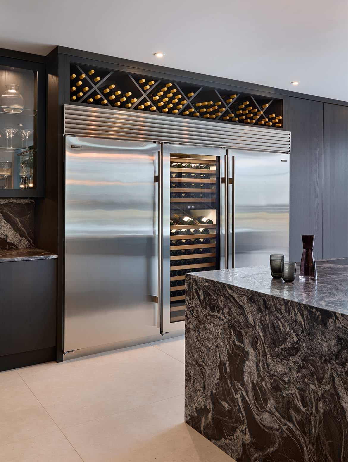 A modern kitchen in Black. The edge of the granite central island oversees the bespoke cabinetry surrounding Sub-Zero refrigeration. Nestled between the two fridge-freezers, a wine cabinet sits flush. Just visible to right of the refrigeration bank, the push-to-open Valencia doors enclose the double pantry. The bespoke cabinetry extends to the wine storage above the Sub-Zeros while the glass-panelled cupboards reveal shelves of wine glasses within. 