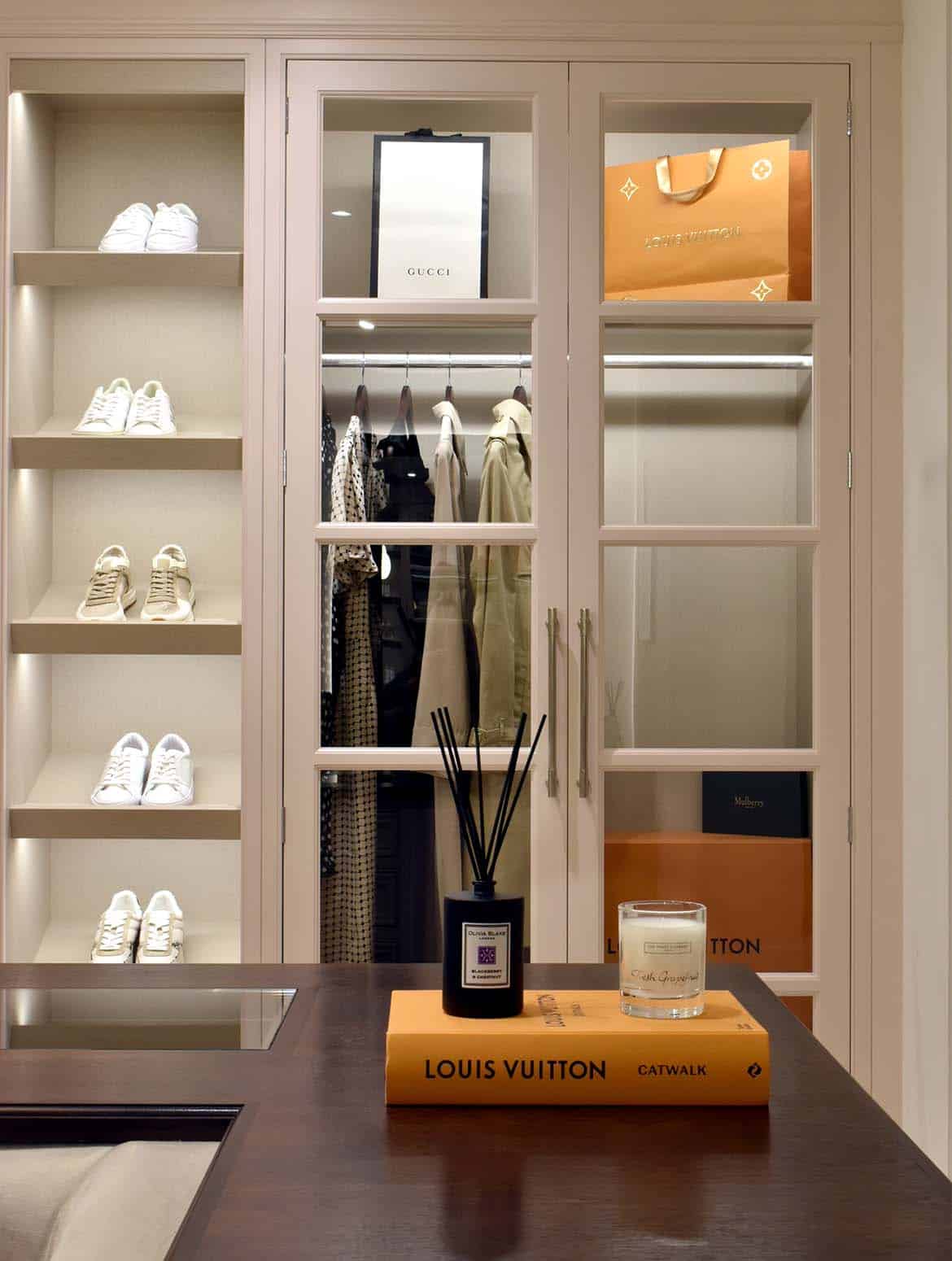 A bespoke modern dressing room interior. The right side of the floor-to-ceiling wardrobe has a five shelves of trainers. To it’s left, there’s a shelf above a close rail enclosed within glass-fronted doors. The central sofa-cum-storage unit foregrounds this view and on the right arm of its U-shape a Louis Vuitton picture book lays beneath an incense and a candle. 