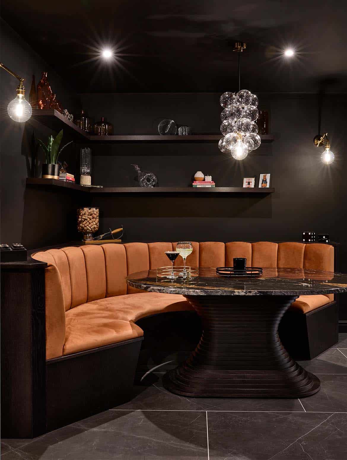 A bespoke bar area in onyx. The orange velvet booth set into the bespoke cabinetry melds easily with the Off-Black walls around it. The Orinoco Granite table reflects light from the chandelier above along with the two cocktails on the table. The shelving above moulds around the corner and has a selection of books, memorabilia, ceramic pots, and plants. 
