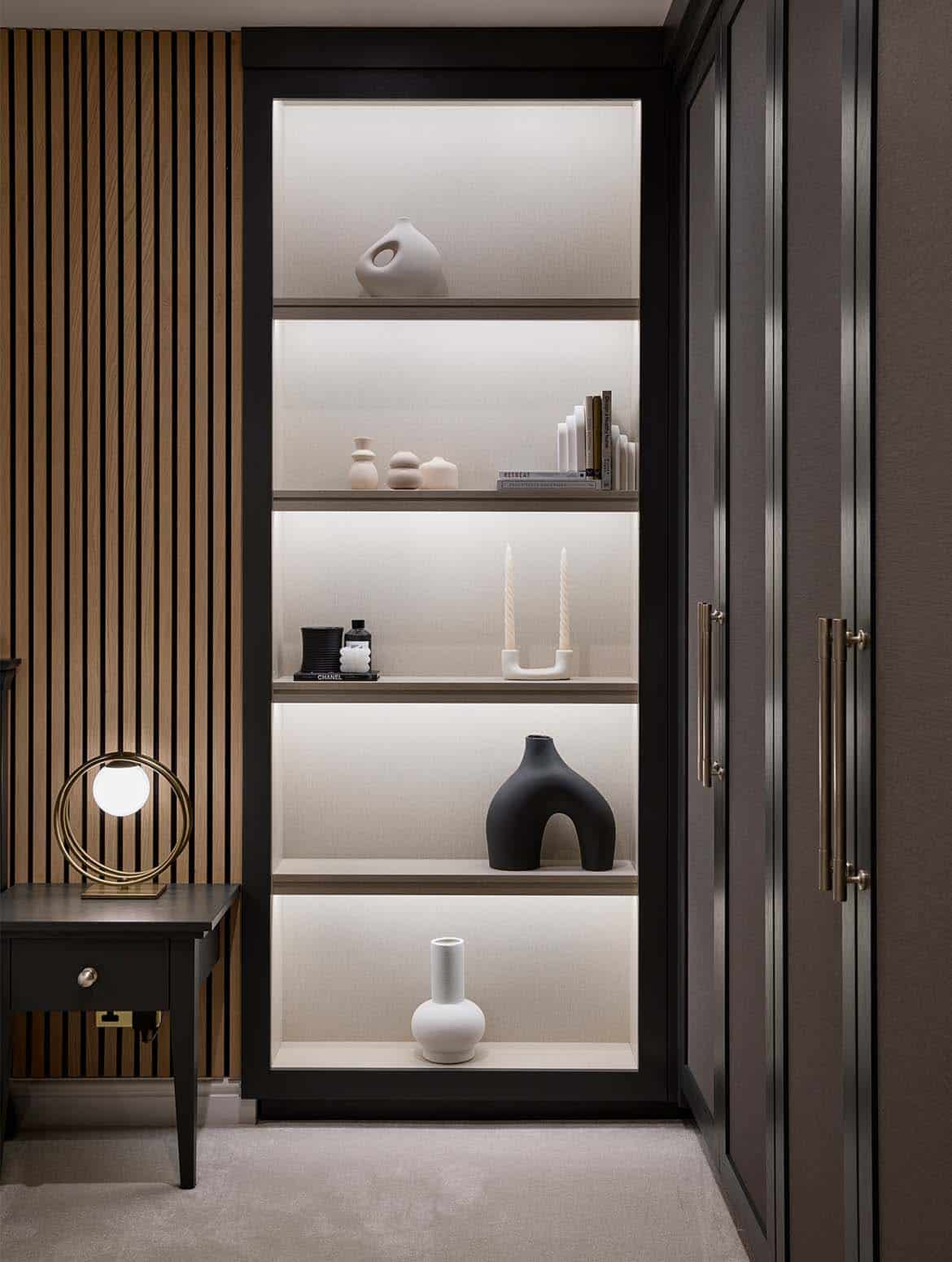 A bespoke modern bedroom interior. The bespoke floor-to-ceiling wardrobe doors guide the eye to well-lit shelves where a mixture of ceramic urns, sculptures, and vases adorn them. To their left, a brown beside table with a circular lamp backs on to the bespoke panelling of the wall behind the bed board.   