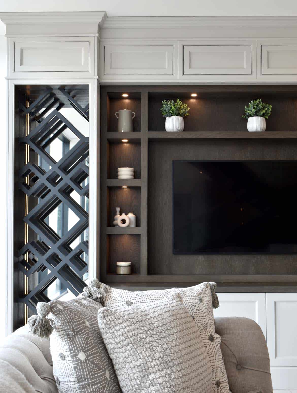 A bespoke media wall in French Grey. The cushions of the Tetrad sofa overlook the bespoke media wall in French Grey. Though blurry and in the background, the mounted television forms the focal point for the cabinetry and the Edwardian panelling goes well with the shelving around the unit that house ceramic urns and jugs. 