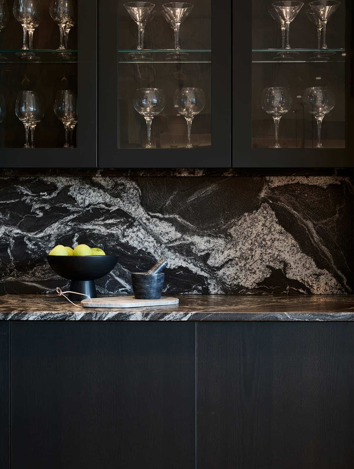 A modern kitchen in Black. The bespoke cabinetry of the cupboards above and below are glazed to ensure a seamless aesthetic. On the worktop, a bowl of apples and chopping board are overseen by the glass-panelled cupboards above. A selection of different wine and cocktail glasses line the shelves. 