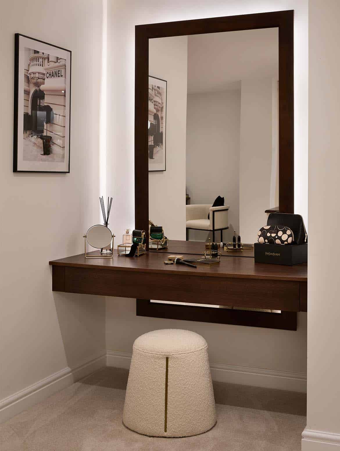 A bespoke modern dressing room interior. A vanity unit held between two walls stands above a small, cone-like stool. On the table, there’s a selection of perfumes, makeup, and a small mirror. A large brown-framed mirror is set against the wall and it’s backlit. On the left hand wall, a framed photo of a Chanel outlet oversees the table. Reflected in the mirror, a chair, upholstered in a similar off-grey colour to the stool can be seen. 