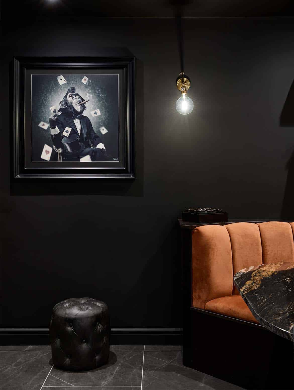 A bespoke bar area in onyx. The edge of the orange velvet booth seat and its accompanying Orinoco Granite table guide the eye to the artwork on the Off-Black wall. The chimpanzee wearing a tuxedo smokes a cigar while watching playing cards scatter around him. Below the frame a black footstool reflects light from the expose bulb above. 
