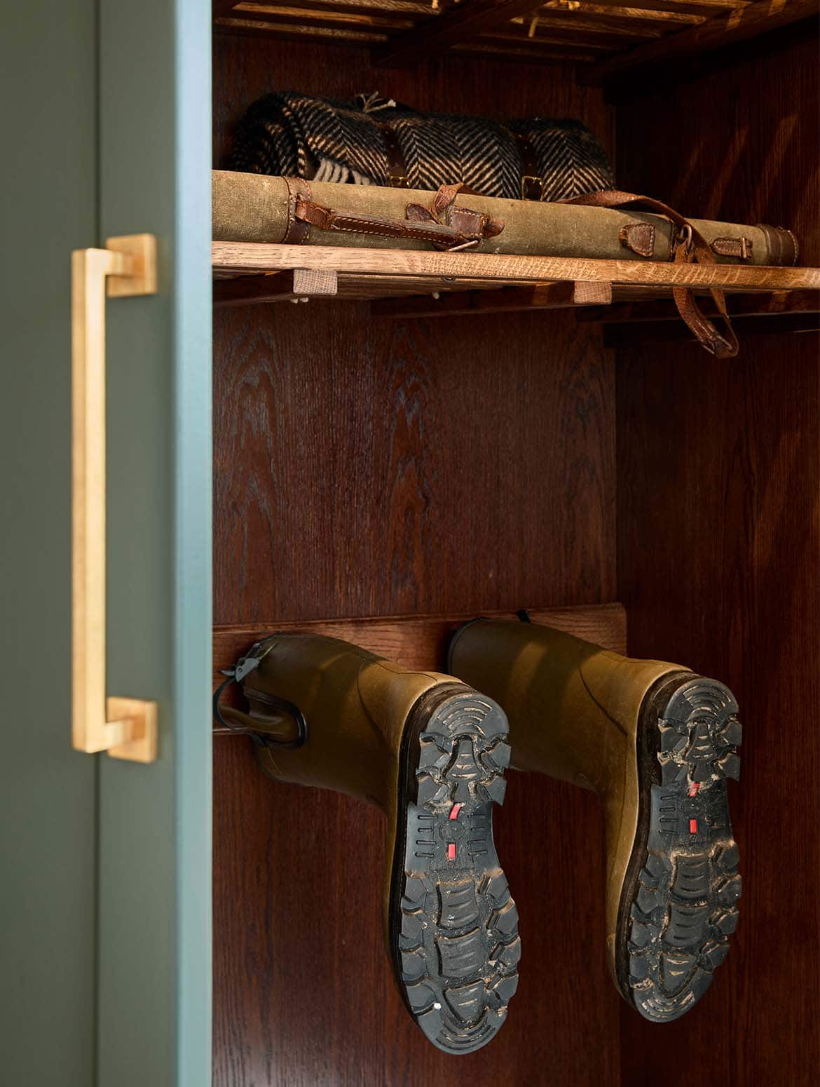 A bespoke bootroom in Studio Green. The open door-with a handle from Armac Martin’s Bromsgrove collection-of the floor-to-ceiling cabinetry reveals bespoke shelving. On the shelf visible, folded and bundled picnic blankets stand in shadow, while below a boot rack houses a pair of wellington boots. 