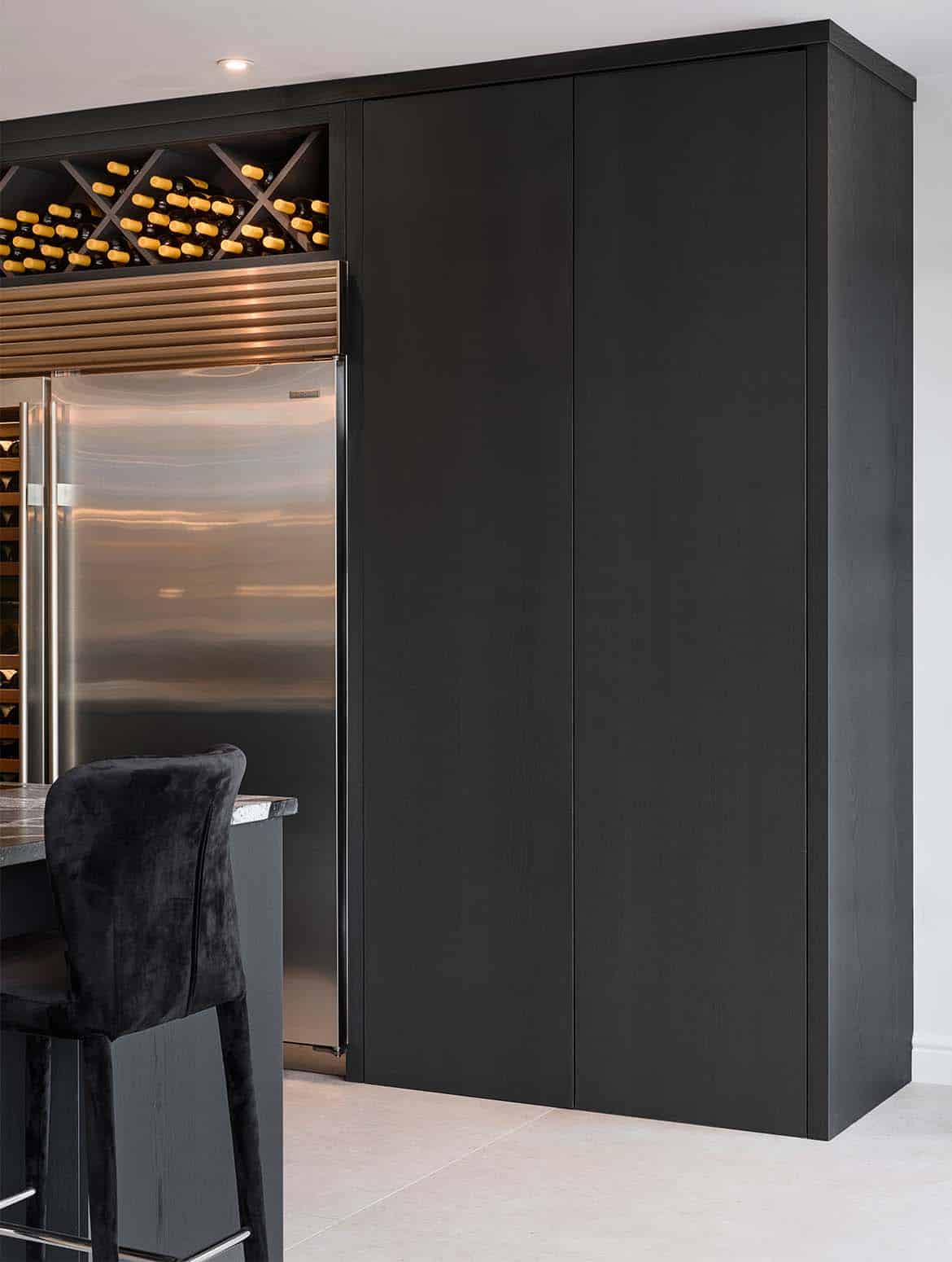 A modern kitchen in Black. The edge of the granite central island oversees the bespoke cabinetry surrounding Sub-Zero refrigeration. The Valencia door of the floor-to-ceiling double pantry blends seamlessly with the cabinetry beyond. Just visible above the Sub-Zero, bespoke wine shelving houses numerous bottles. 