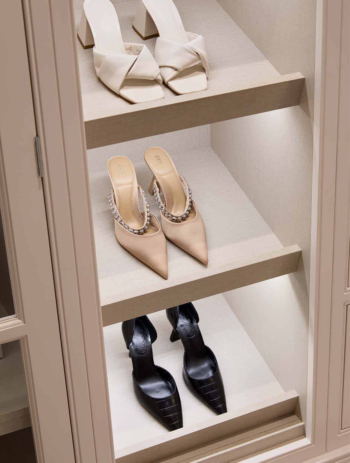 A bespoke modern dressing room interior. A close in view of the underside-lit shoe shelves. There are three of the lower shelves visible and on each, a pair high heels face out to the dressing room at large. They’re surrounded by the bespoke Edwardian profiling of the cabinetry painted in Shadow White. 