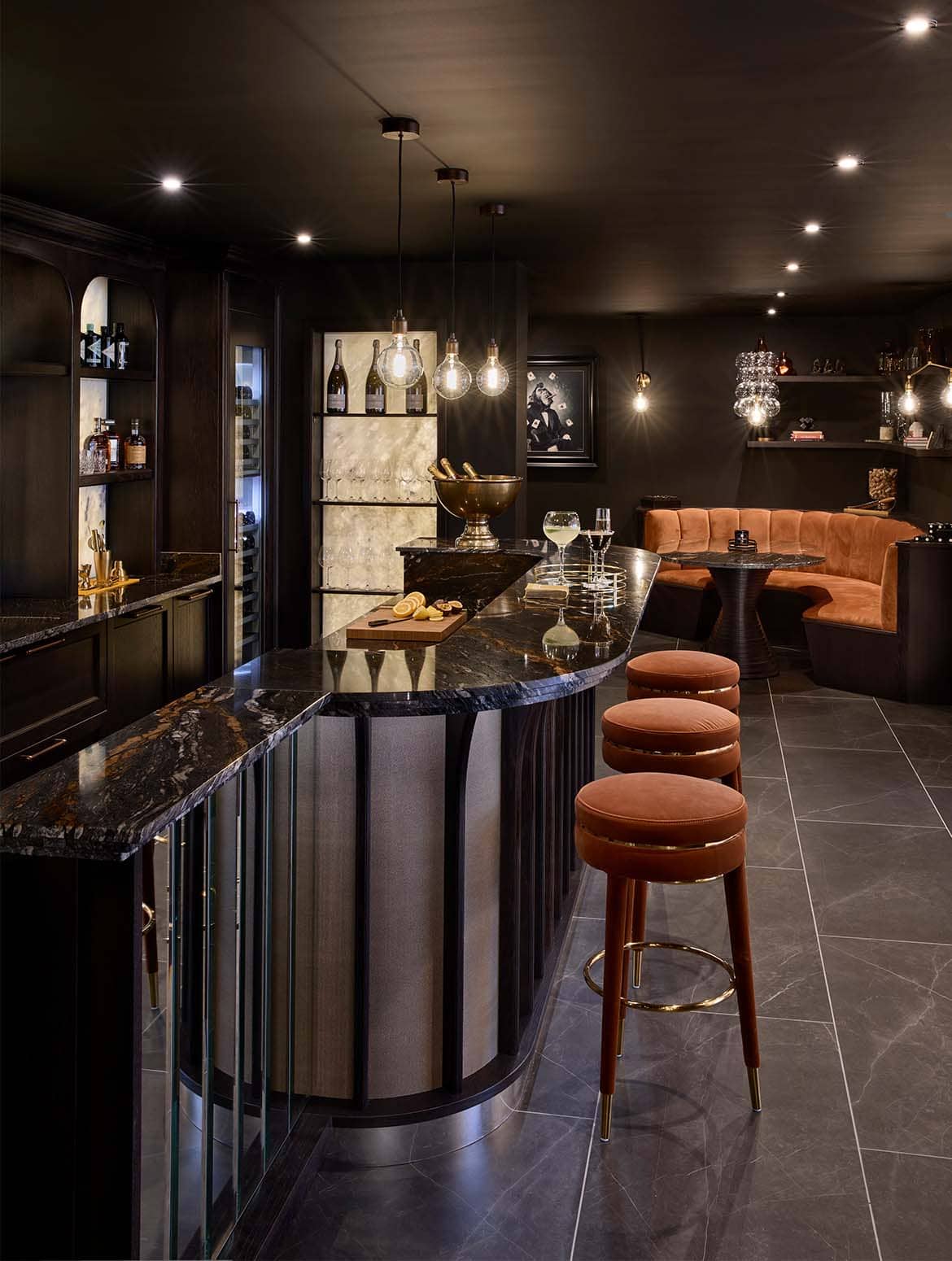 A bespoke bar area in onyx. The well-lit orange velvet upholstered booth catches the eye ahead, while upholstered stools in matching velvet stand before a Orinoco Granite countertop. An assortment of drinks stand in a tray on the bar countertop, while behind garnishes for drinks like lemons and orange lie sliced on a chopping board. On the bespoke cabinetry behind the bar, a built in wine fridge reflects light from the exposed bulbs above. 