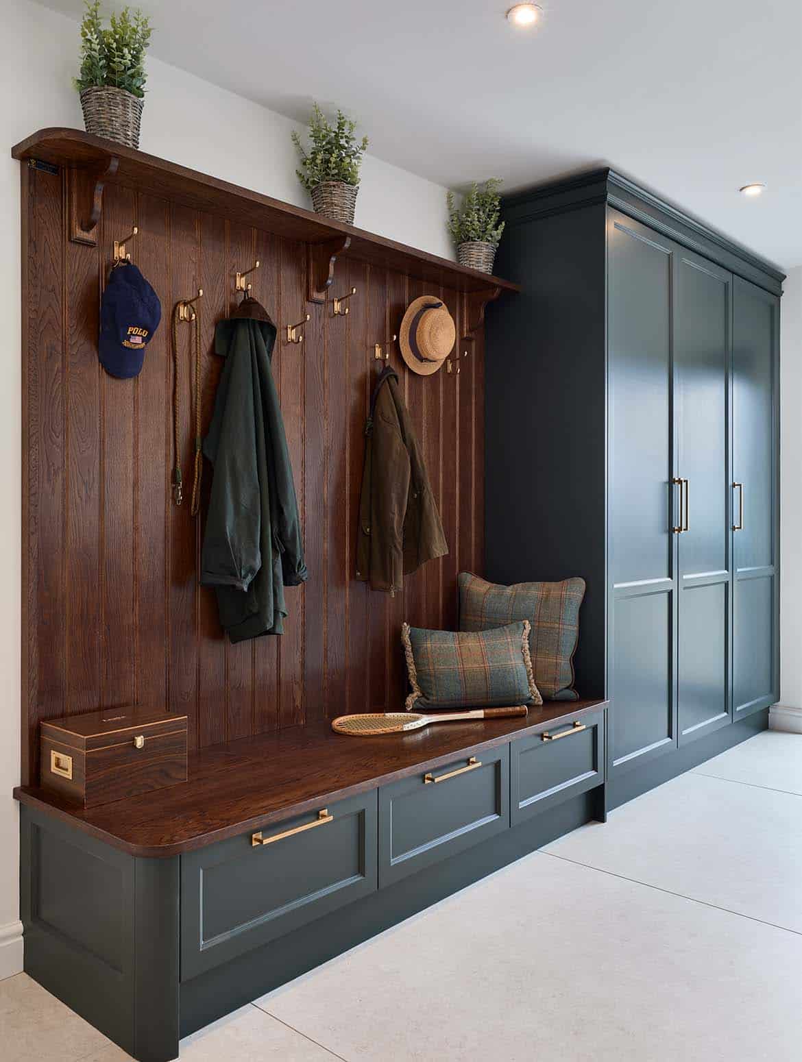 A bespoke bootroom in Studio Green. A crafted coat store that melds seamlessly into the floor-to-ceiling cabinetry beyond. Three green plants in wicker baskets spaced equally across the top line of the clothes storage appear to oversee the clothes hooks with two jackets and two hats hung up below. A footlocker and an old-fashioned tennis racket sit on the ledge that allows for people to sit to put their shoes on. There are three drawers underneath this ledge. The doors to the cabinetry beyond are similarly handled—in handles from Armac Martin’s Bromsgrove collection. 