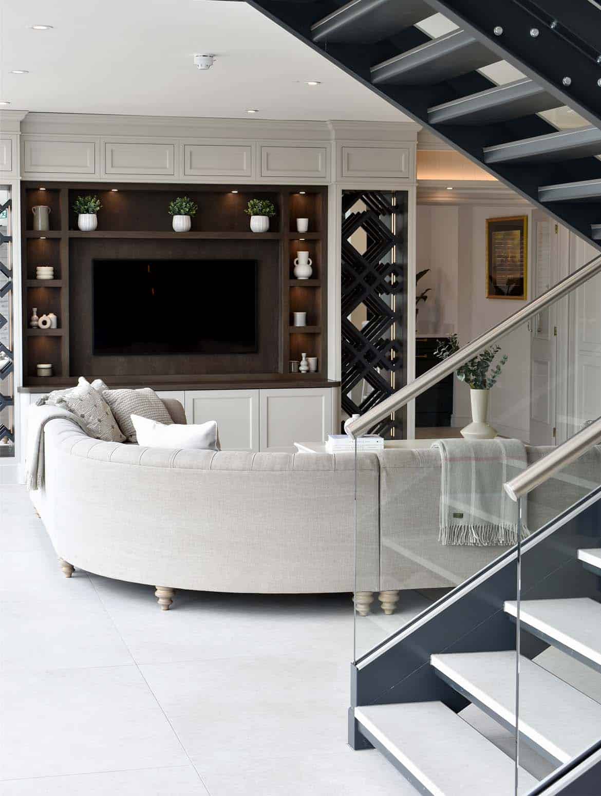 A bespoke media wall in French Grey. A staircase frames the view towards the media wall. The mounted television, focal point for the wall, oversees a Tetrad sofa and a coffee table with a ceramic vase on its surface. Surrounding the television, bespoke shelving, set into floor-to-ceiling cabinetry, adds charm through its assortment of ceramic urns and vases, complementing the diagonal woodwork of the main pillars to the cabinet. 