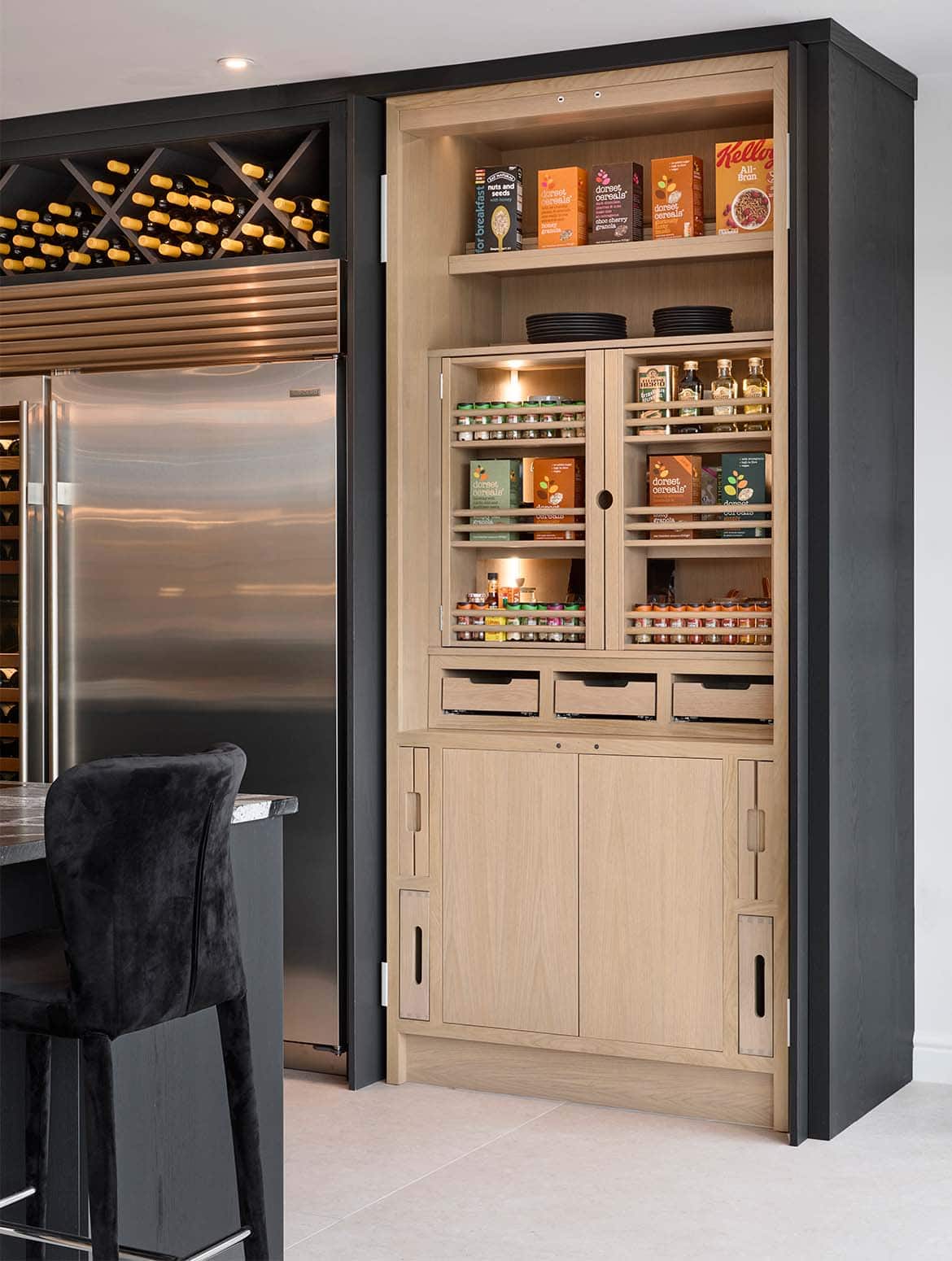 A modern kitchen in Black. The edge of the granite central island oversees the bespoke cabinetry surrounding Sub-Zero refrigeration. The open Valencia doors of the floor-to-ceiling double pantry blends seamlessly with the cabinetry beyond. Inside, the oak satin finish of the shelving and the drawers stands out well. At the top of the pantry, cereal boxes line the shelf.  