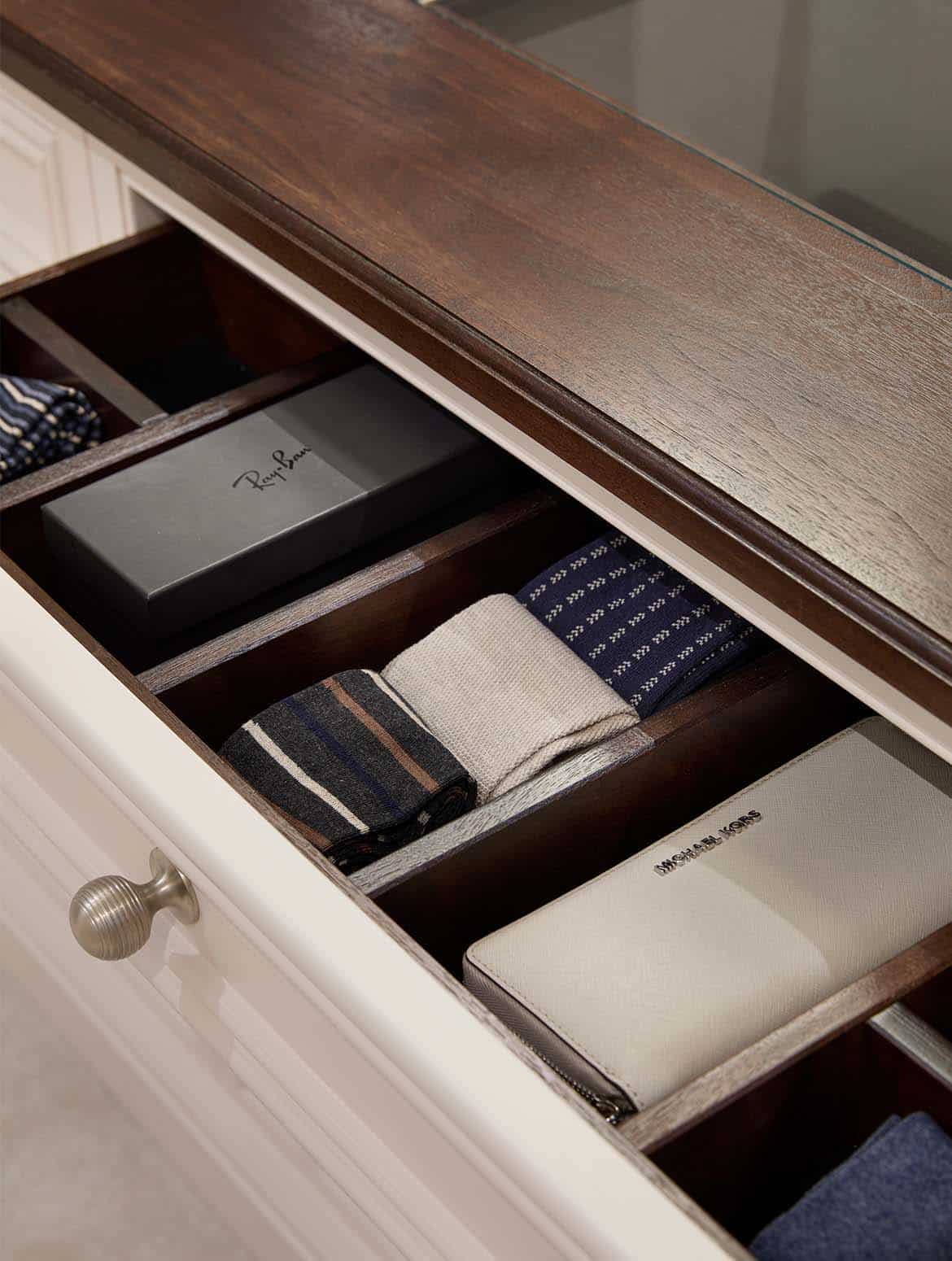 A bespoke modern dressing room interior. A close in view of the top drawer of middle bespoke sofa-cum-storage cabinetry. Fitted with Armac Martin handles, the drawer has a mixture of socks, wallet, and Ray Ban sun glasses. The Shadow White of the Englemere panels offsets the walnut finish of the cabinetry’s top. It’s just visible that a pane of glass on the surface gives a bird’s eye as to what’s inside the drawer when it’s closed. 