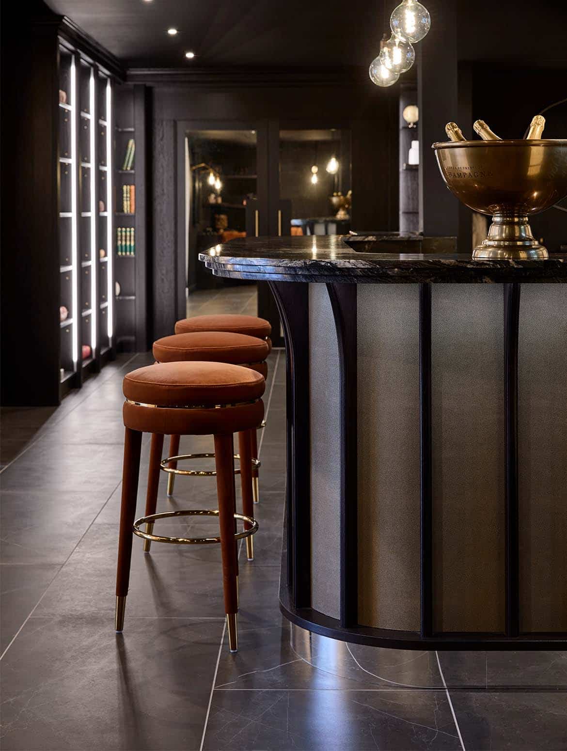 A bespoke bar area in onyx. The upholstered stools in orange velvet catch the eye stood before a Orinoco Granite countertop. They guide the eye towards the library shelves beyond. On the countertop, a bucket with three champagne bottles catches light from the exposed bulbs above.   