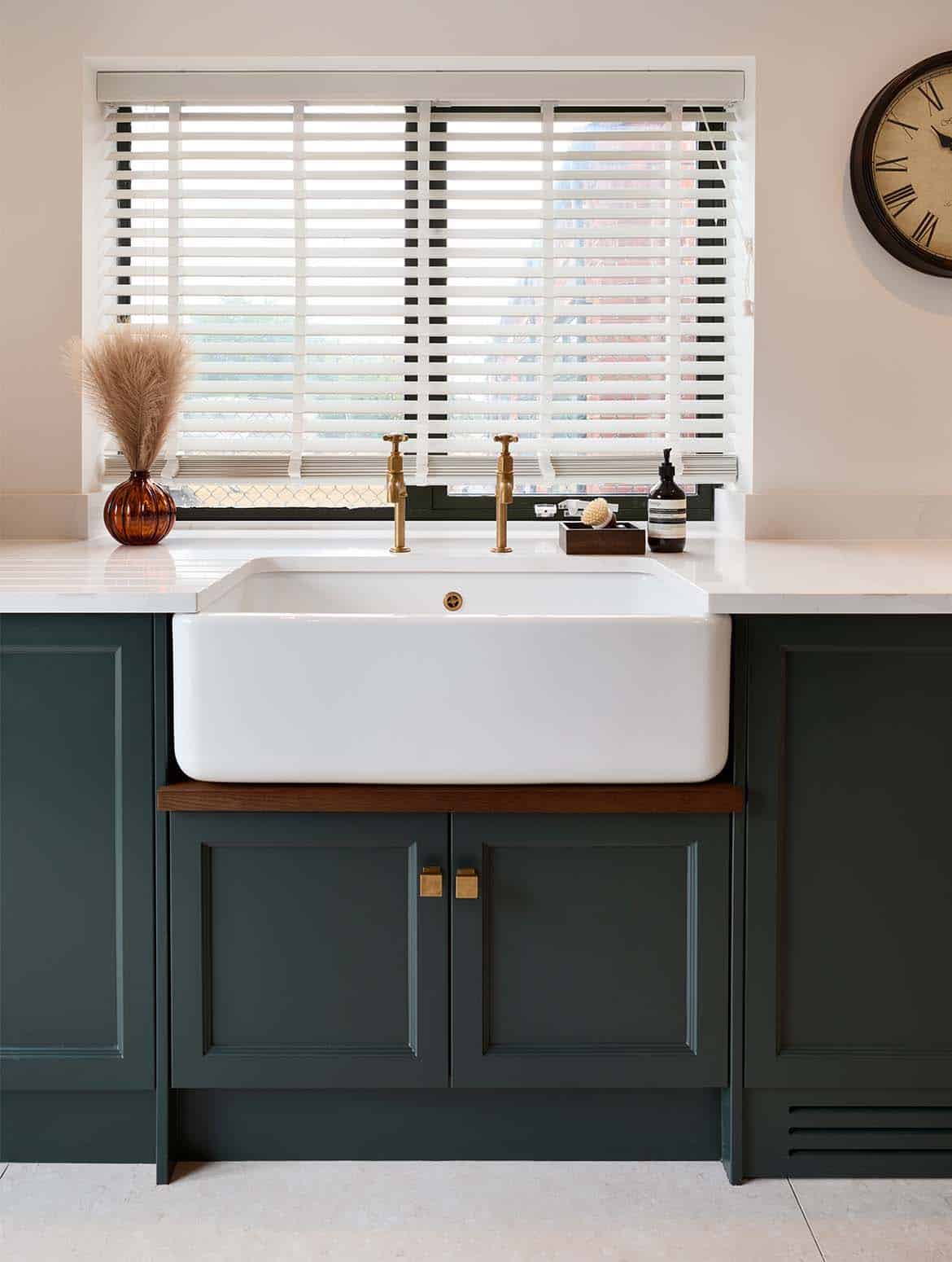 A bespoke bootroom in Studio Green. A view of the Belfast sink and burnished taps set into the white worktop. Next to the cold tap, a small tray with a brush and hand soap stands opposite a small ornamental glass vase. The bespoke cabinetry below the sink contrasts well with the white worktop above. The handles, taken from Armac Martin’s Bromsgrove collection, on the cupboards below contrasts well with the Studio Green panel colour and the similarly burnished taps above.  