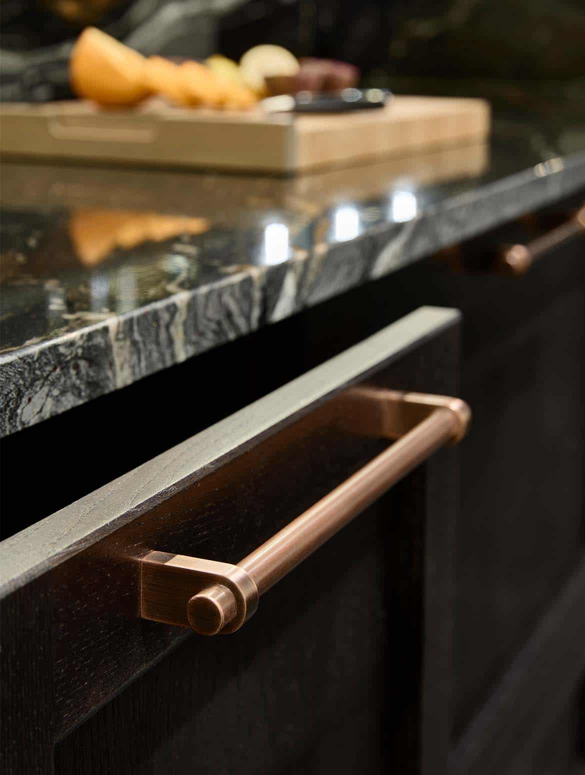 A bespoke bar area in onyx. A close in view of an open drawer beneath the Orinoco Granite countertop. The stained vintage tobacco on ash colouration of the drawer contrasts its burnished bronze handle well. Though out of focus, sliced orange and lemons are visible on a chopping board on the counter behind. 