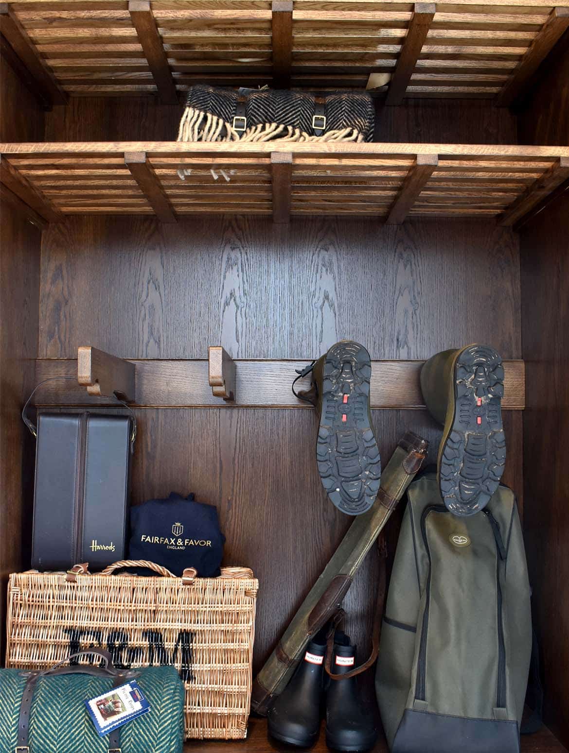 A bespoke bootroom in Studio Green. A view of the open storage cabinet with bespoke shelving. The top shelves have folded and bundled picnic blankets in shadow, while below a boot rack houses a pair of wellington boots. Beneath the boots, a Fortnum & Mason hamper along with another folded blanket and wellington boots fill the space at the foot of the cabinet.  