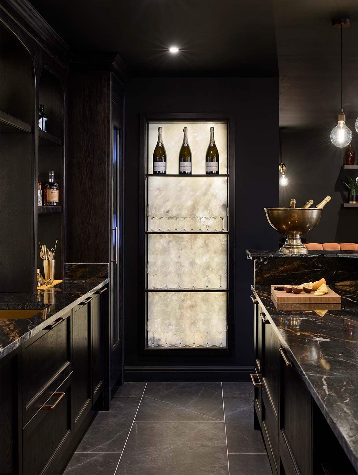 A bespoke bar area in onyx. The Orinoco Granite surfaces of the bespoke cabinetry against the wall and behind the bar guide the eye towards the backlit onyx backsplash behind the glass shelves ahead. On the bar countertop a chopping board with slices of orange and pomegranate   lie opposite the visible cocktail shaking on the back wall. They almost steal attention away from the three magnums of champagne on the top shelf ahead.  