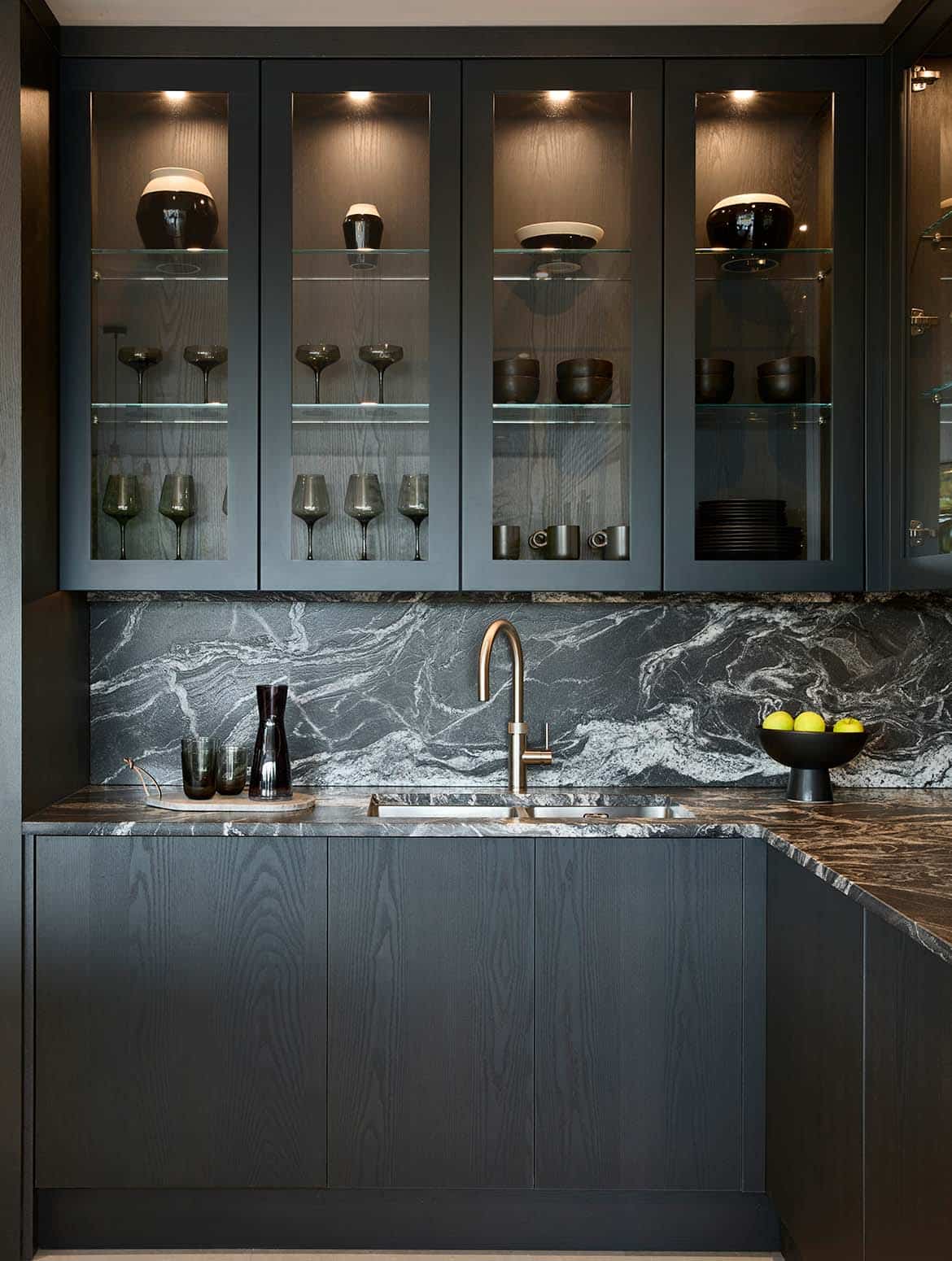 A modern kitchen in Black. The right-angle of the bespoke cabinetry contrasts the worktop and Quooker tap set into it. The glass-panelled cupboards above reveal a selection of glasses and ceramic urns. The Valencia panelling of the cupboard doors blends seamlessly with the cabinetry.