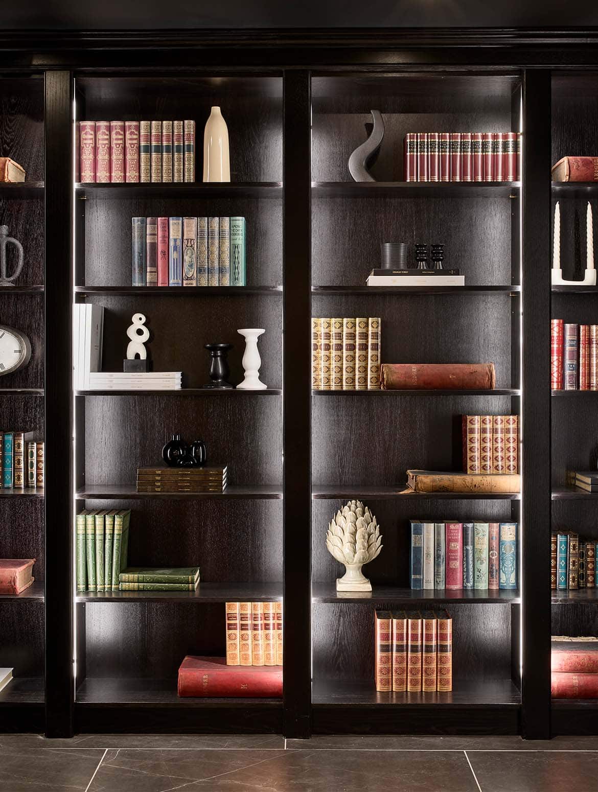 A bespoke bar area in onyx. The four off-black library shelves hold an assortment of ornaments, books, and tomes on their side. The shelves have a ghostly quality with the inbuilt light around their periphery. 