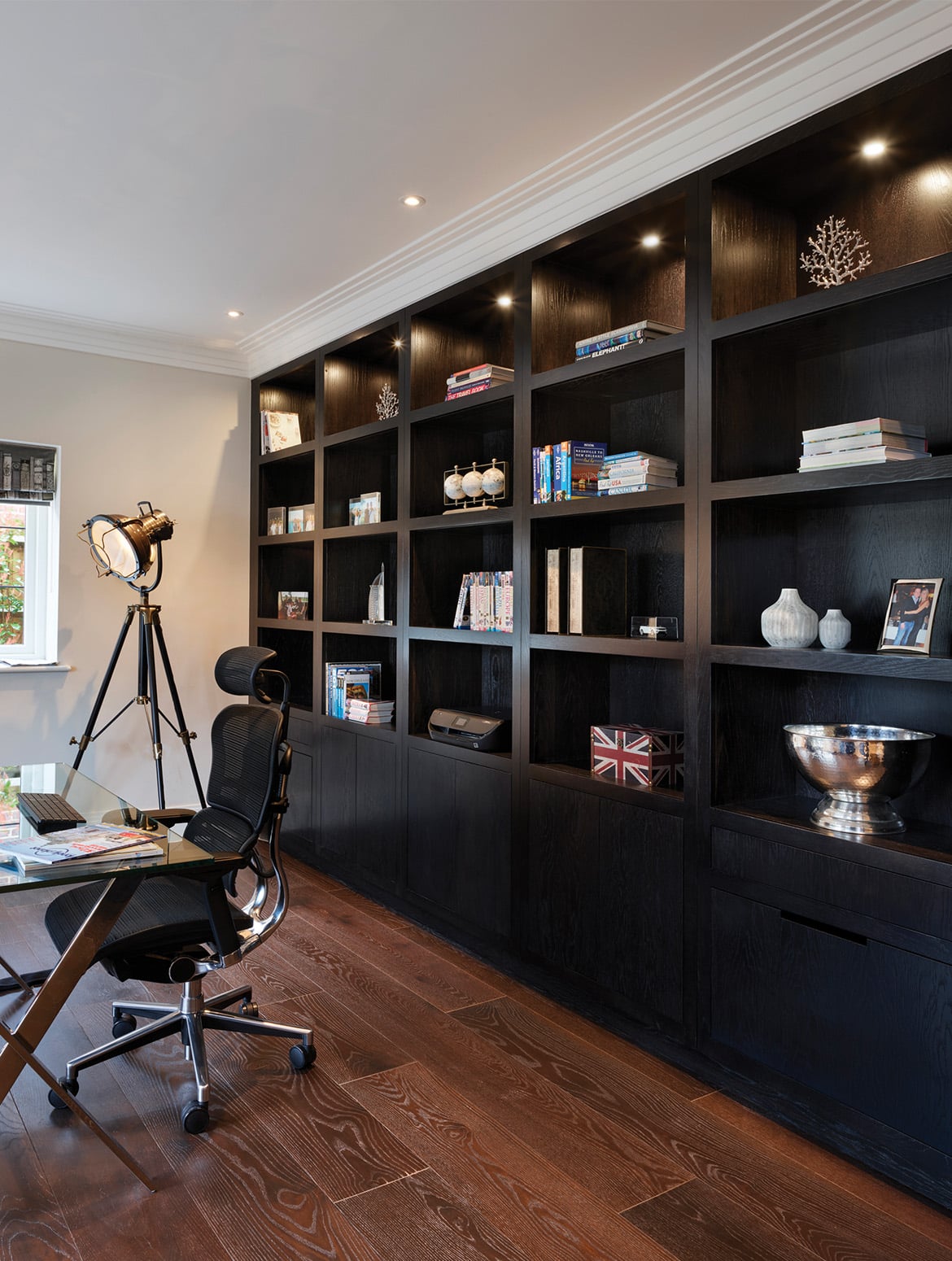 A home office with floor-to-ceiling bespoke cabinetry. The black wood stretching the length of the room has cupboards along the first level and then exposed shelving for the next four. The shelves are adorned with a variety of items ranging from a bespoke urn, memorabilia, books, and a set of three small globes. In the far corner, an old fashioned film set light looks over the modern glass desk with ergonomic desk chair sat underneath it. 