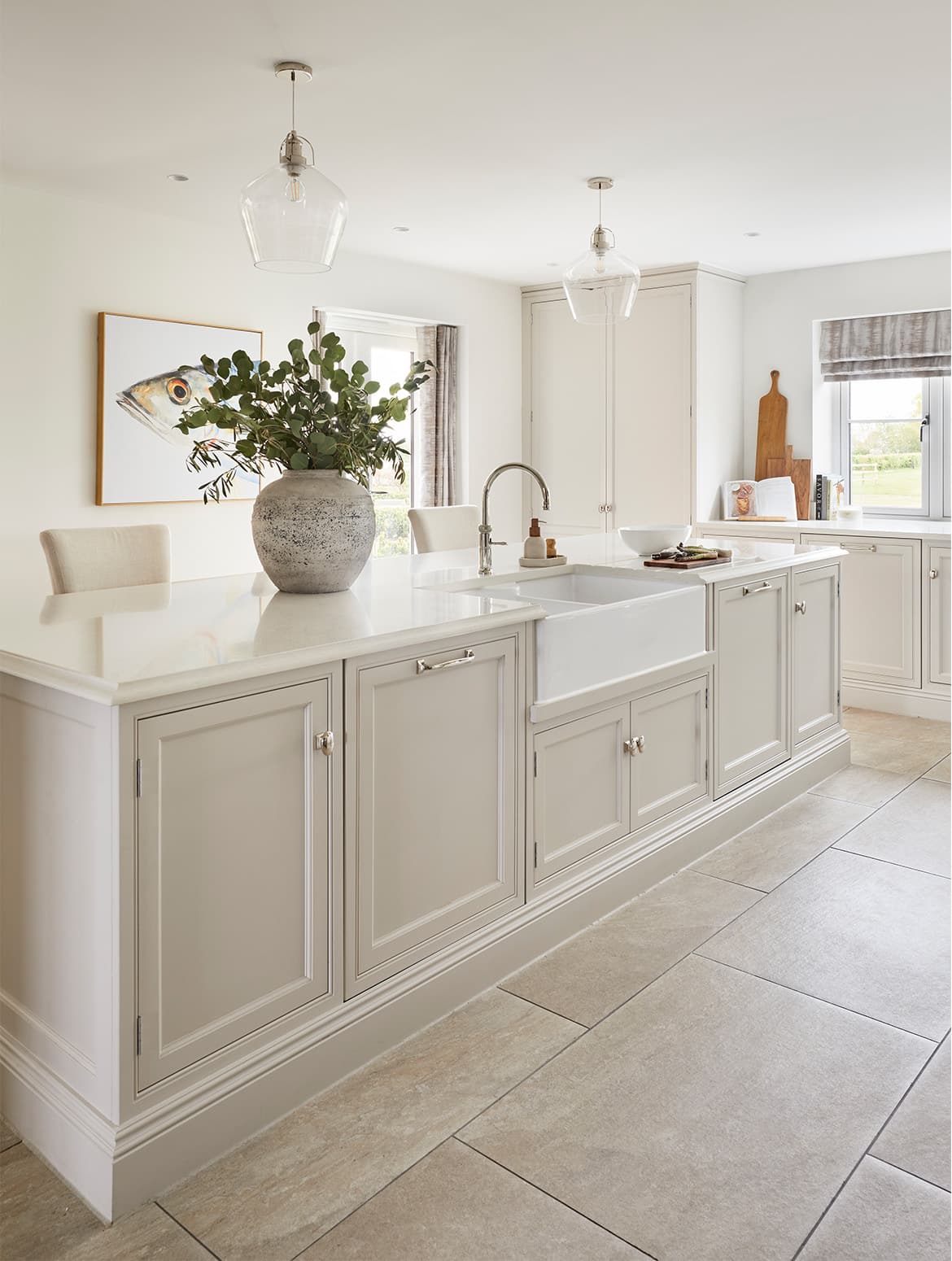 A traditional shaker kitchen in French Grey. The white central island has a Quooker sink and a large ceramic vase on it. On the draining board bevelled in to the worktop, there is a ceramic bowl and chopping board. Just beyond, a floor-to-ceiling panty stands flush to cabinetry against the wall underneath a window. 
