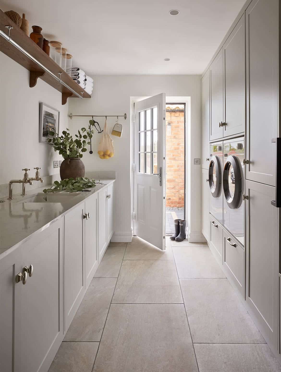 A bespoke bootroom in French Grey. The bespoke cabinetry fitted with handles from Armac Martin’s Bourneville’s collection guides the eye towards the ajar utility door. Nestled within the cabinetry, a tumble dryer and a washing machine oversee the similarly crafted cupboards and worktop, fitted with Belfast sink. Above the sink, a towel-laden shelf oversees the rail by the door. The rail has a stethoscope, a net of lemons, and a brush hanging from it. A pair of black wellington boots sit in the open doorway and a large ceramic pot stands on the worktop.