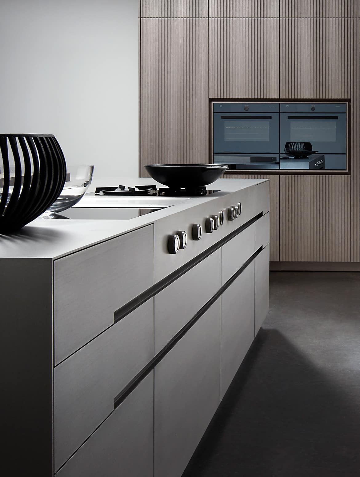 A modern kitchen in smoked wood finish. The floating island stands central to bespoke cabinetry against the back wall. The bespoke cabinetry surrounds two V-Zug single ovens and these oversee the built-in hob and burner. Flush handleless drawers ensure the sleek modern finish to the island.    