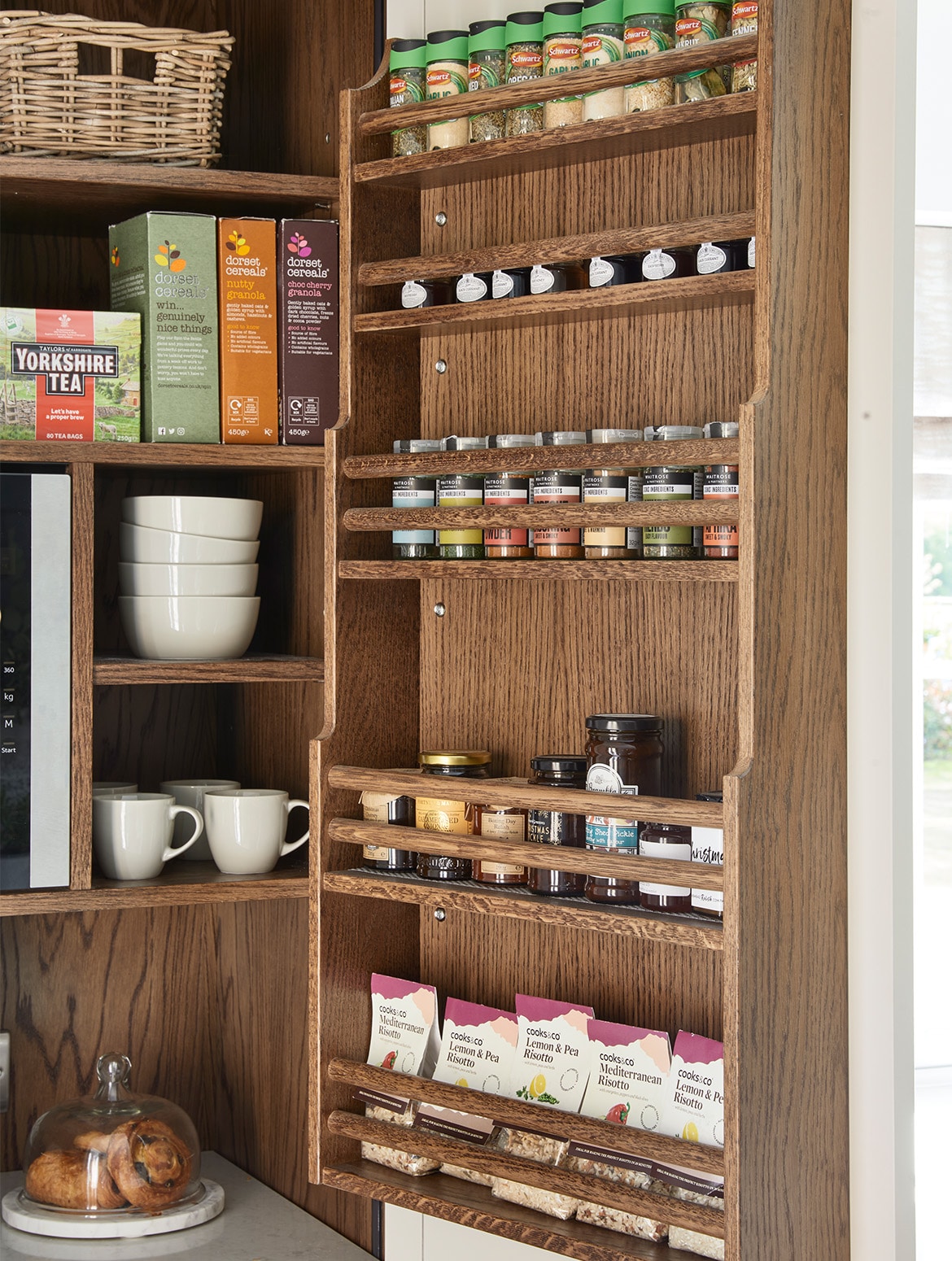 A traditional shaker kitchen in French Grey. The open door of a floor-to-ceiling pantry contains a selection of spices opposite shelves that have breakfast items and breakfast pastries too.