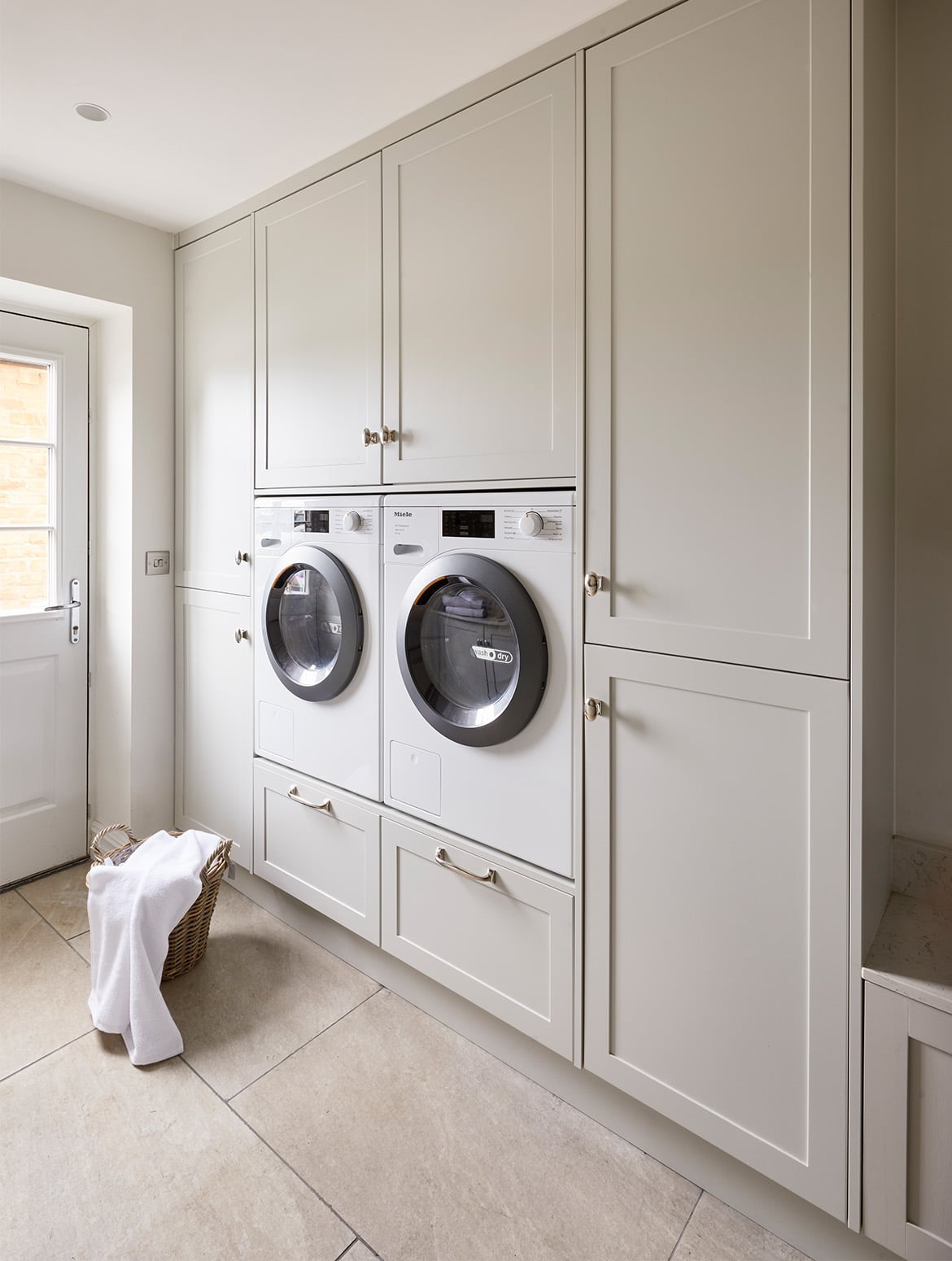 A bespoke bootroom in French Grey. The bespoke cabinetry fitted with handles from Armac Martin’s Bourneville collection houses a tumble dryer and a washing machine. The floor-to-ceiling cabinetry covers the back wall, while the white utility door is just visible to the far left. On the floor in front of the cabinets, a wicker basket has a white towel laid over it. 