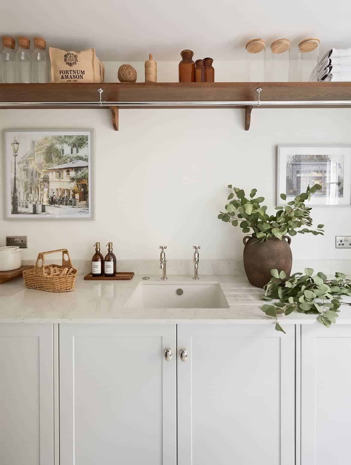 A bespoke bootroom in French Grey. The bespoke cabinetry fitted with handles from Armac Martin’s Bourneville collection catches the eye, while the worktop above accommodates a ceramic vase next to the taps and hand soap to its left. Two framed pictures of Paris hold the left and the right side of the wall behind the worktop. Above, a laden shelf has empty glass jars, a Fortnum & Mason bag, a neatly-stacked pile of towels on it. 