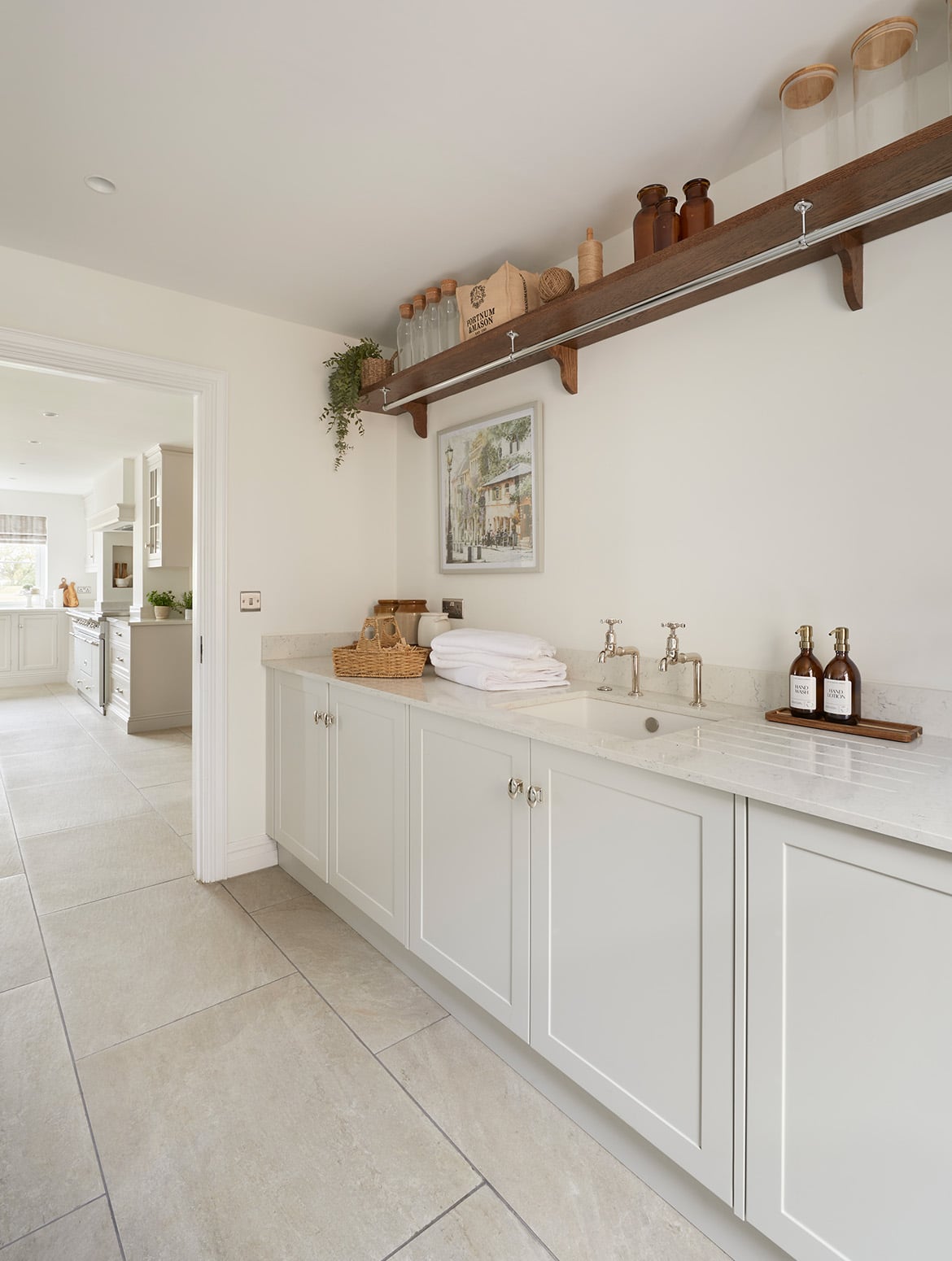 A bespoke bootroom in French Grey. The bespoke cabinetry fitted with handles from Armac Martin’s Bourneville collection catches the eye, while the worktop above accommodates a ceramic vase next to the taps and hand soap to its left. A framed pictures of Paris holds the left side of the wall behind the worktop. Above, a laden shelf has empty glass jars, a Fortnum & Mason bag, and a neatly-stacked pile of towels on it. Beyond the cabinetry, the open doorway reveals the bespoke traditional shaker kitchen beyond. 