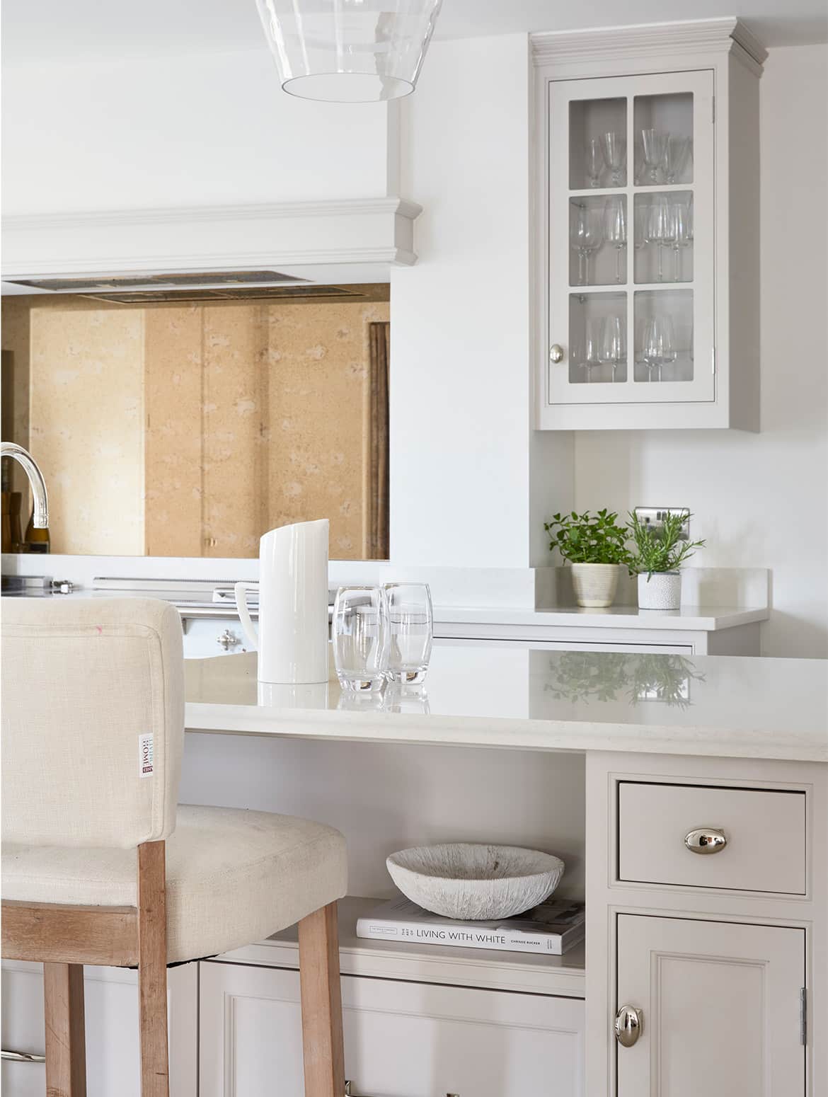 A traditional shaker kitchen in French Grey. The white central island has a ceramic water jug and glasses on it, and has an upholstered high chair underneath a slight lip. Armac Martin handles stand out on the bespoke island cabinetry and beyond the island, more bespoke cabinetry surrounds a white Lacanche range cooker. 