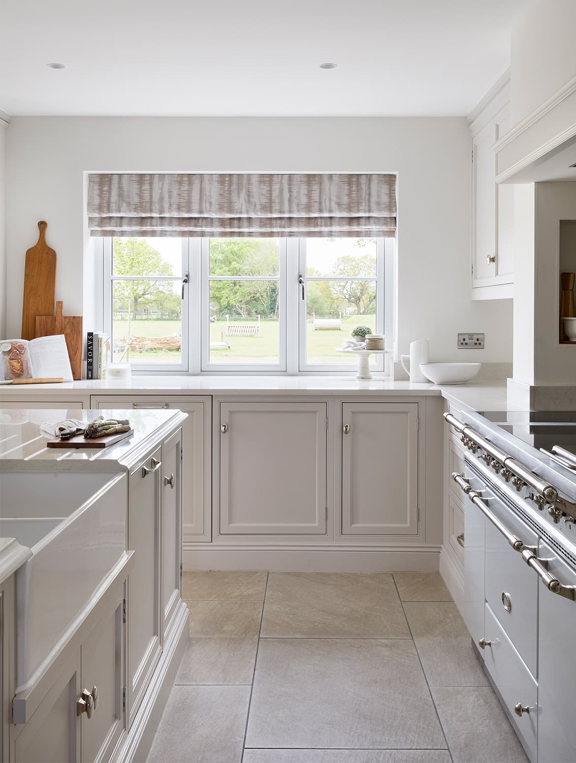A traditional shaker kitchen in French Grey. The view to the garden beyond is guided by the central island on one side and the white Lacanche range cooker on the other. The sink, set in to the island, is visible along with the bespoke draws and cupboards that make up the island. On the side, the Lacanche stands proud to the bespoke work above and along the wall.