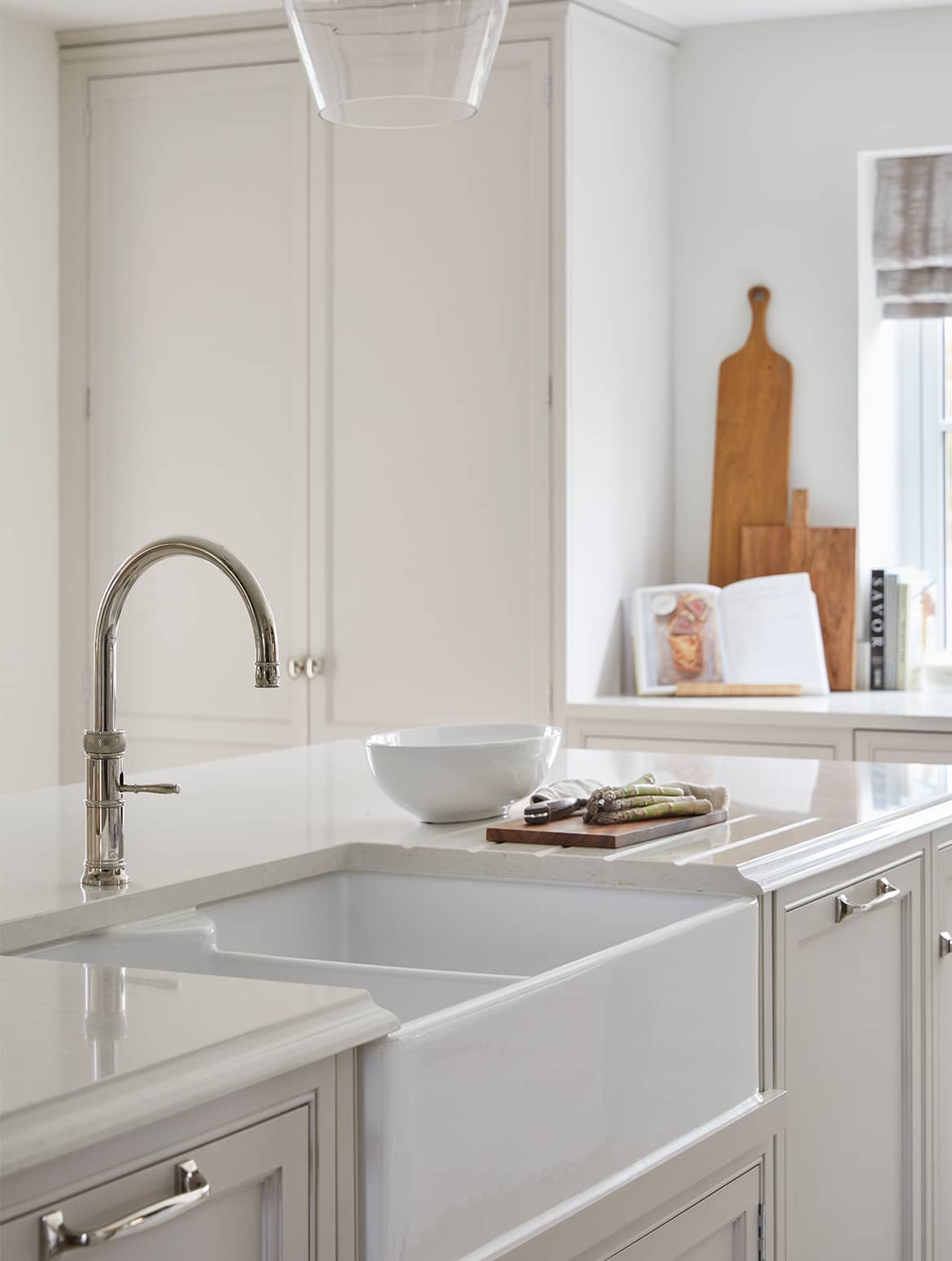 A traditional shaker kitchen in French Grey. The island fitted with Armac Martin handles and fitted with a Quooker tap, stands centrally in the kitchen. A chopping board sits on the draining board bevelled into the marble top. Visible beyond, an open cookbook and chopping boards can be seen.