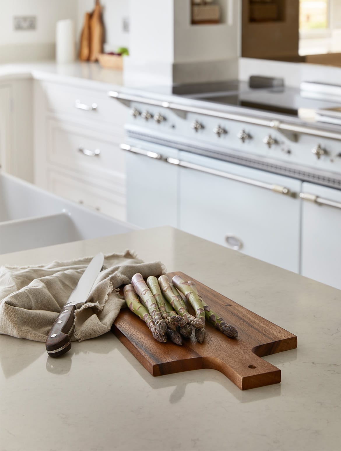 A traditional shaker kitchen in French Grey. On a white central island near the sink, a chopping board with asparagus on it sits ahead of a white Lacanche range cooker set into bespoke cabinetry. 
