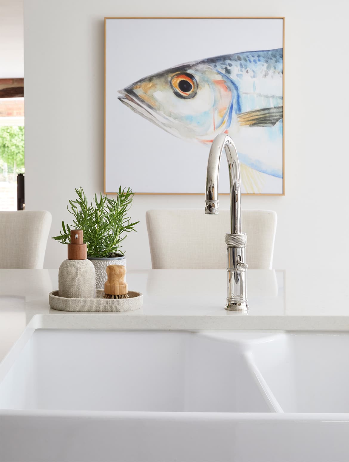 A traditional shaker kitchen in French Grey. The white sink sits centrally, fitted with a stainless steel Quooker tap next to a dish with soap and scrubber. Behind island, upholstered high chairs are visible and also a canvas print on the wall.