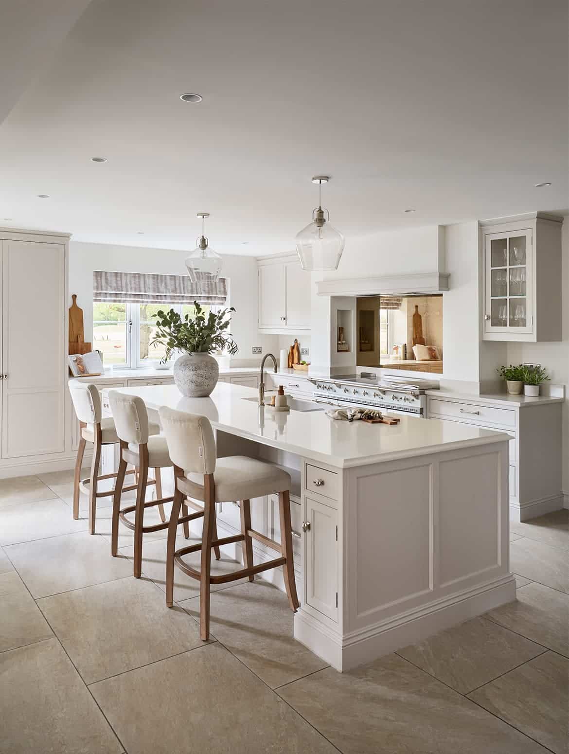 A traditional shaker kitchen in French Grey. The white island fitted with Armac Martin handles and fitted with a Quooker tap, stands centrally in the kitchen. Three upholstered high chairs sit underneath a ledge of the island worktop. Behind the island, bespoke cabinetry stands against the back wall surrounding a white Lacanche range cooker.