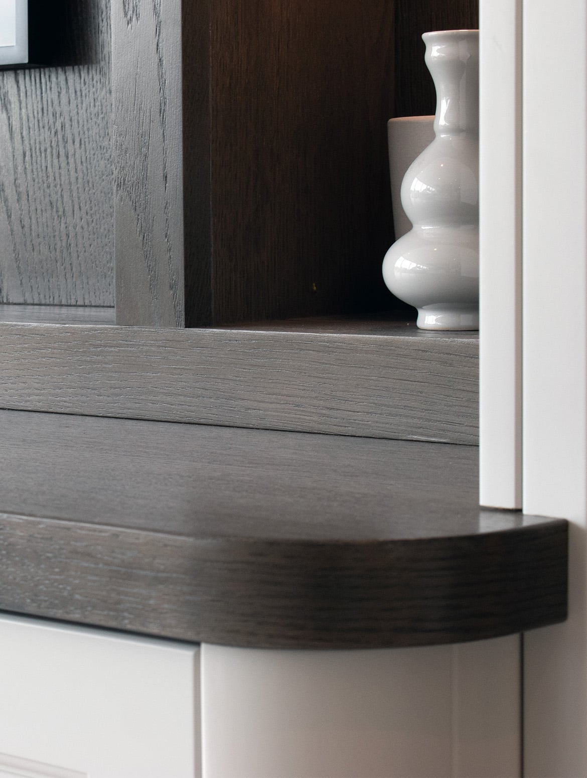 A bespoke media wall in French Grey. The rounded ledge of the cabinet runs out of view, while the French Grey of the bespoke shelving runs upwards in contrast to the darker finish of the rounded ledge. In the shelf of the cabinetry, a similarly grey ceramic vase can be seen reflecting light from its three curves.  