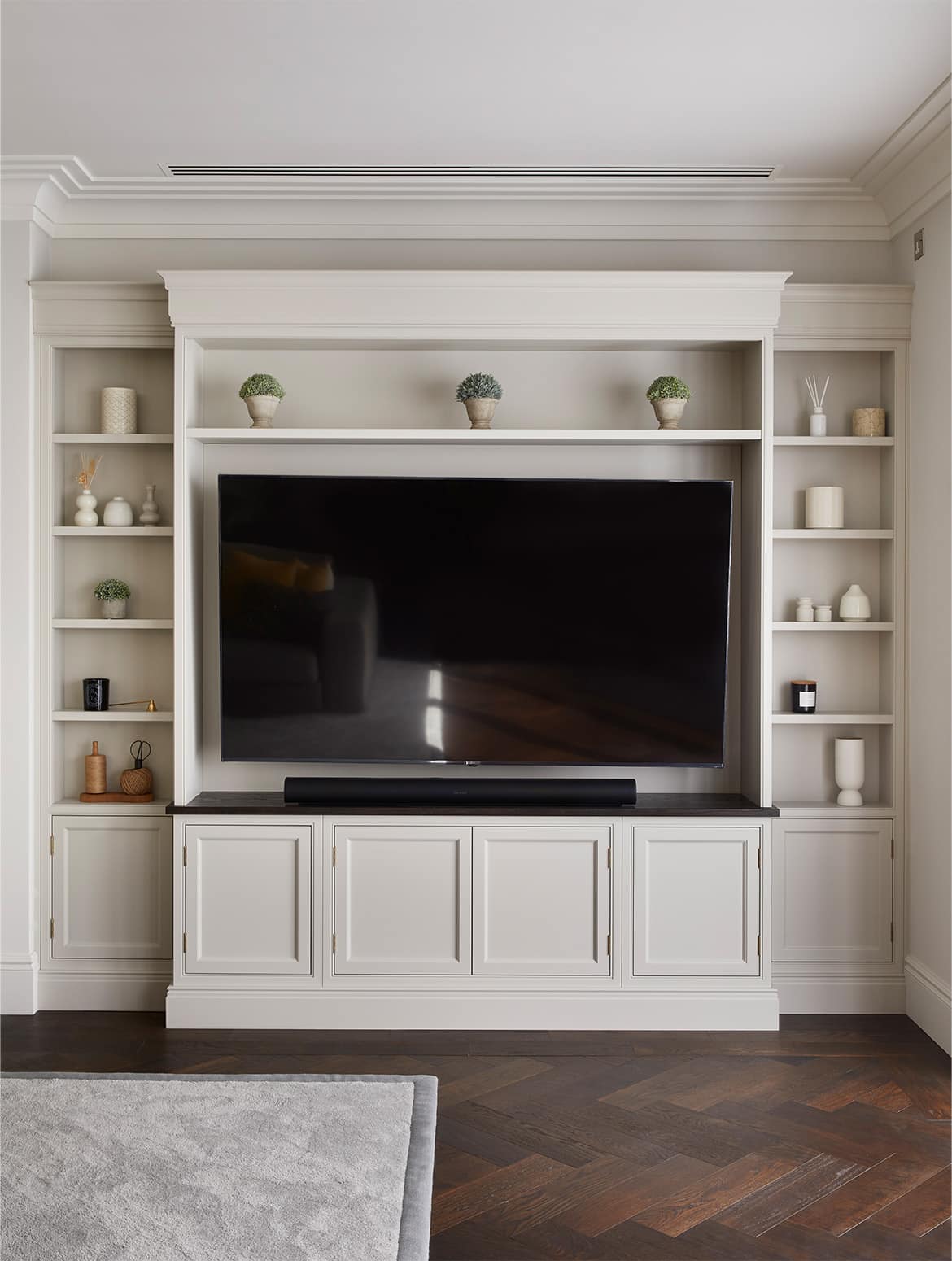A media wall with bespoke cabinetry in French Grey. The right wall has a television mounted in the middle while the surrounding cabinetry has shelves holding an assortment of baskets, ceramic urns, vases and decorative plants. Underneath, push-to-open cupboards mesh seamlessly with the rest of the cabinetry.