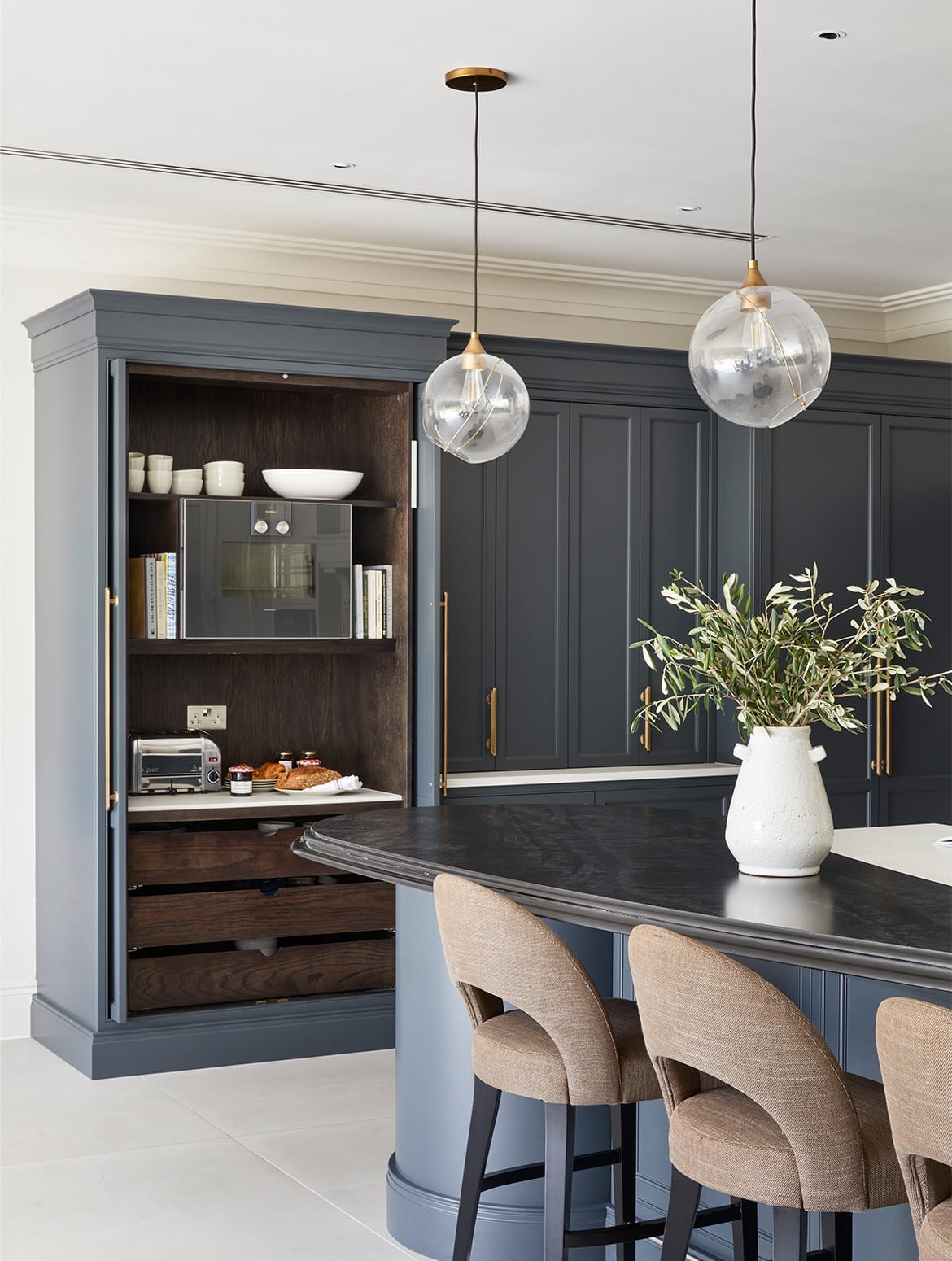 A traditional shaker kitchen project in off-black. An off-black open pantry stands next to the close doors of a hidden pantry. A Gaggenau microwave stands on the second shelf above a selection of breakfast pastries. Breakfast plates and bowls sit on the top shelf as well. This cabinetry overlooks a central island with the bespoke cabinetry and a streamlined wood ledge that contrasts the white worktop of the remaining island beyond. 