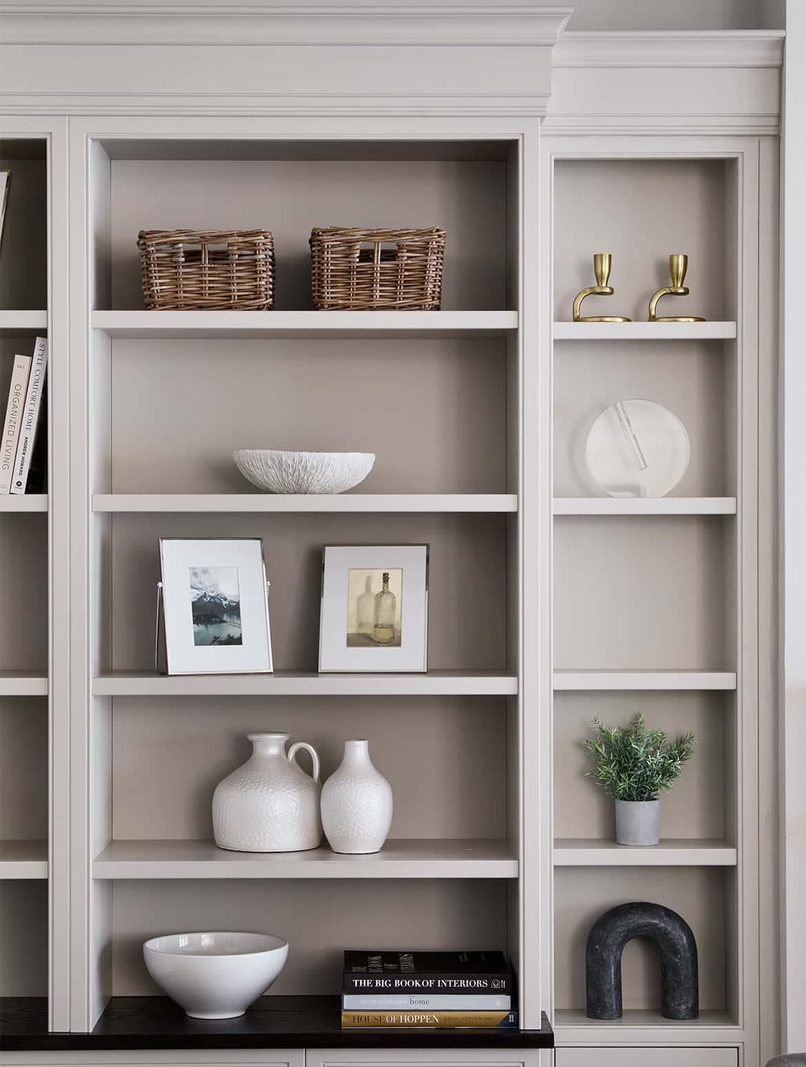 A bespoke media wall in French Grey. A close in view of the shelves of the media wall where a eclectic selection of ornaments, vases, urns, and baskets sit. The chamfered edges of the cabinet top stand proud to the wall behind. 