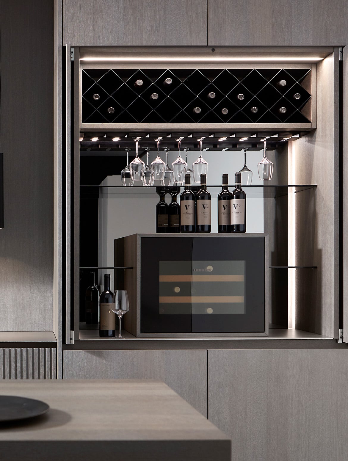 A modern kitchen in smoked wood finish. The open and hidden doors to the bespoke cabinetry reveal a drinks cabinet. Fitted with a built-in wine fridge, the space also boats wine rack and glasses rack set into the underside of the shelf.  
