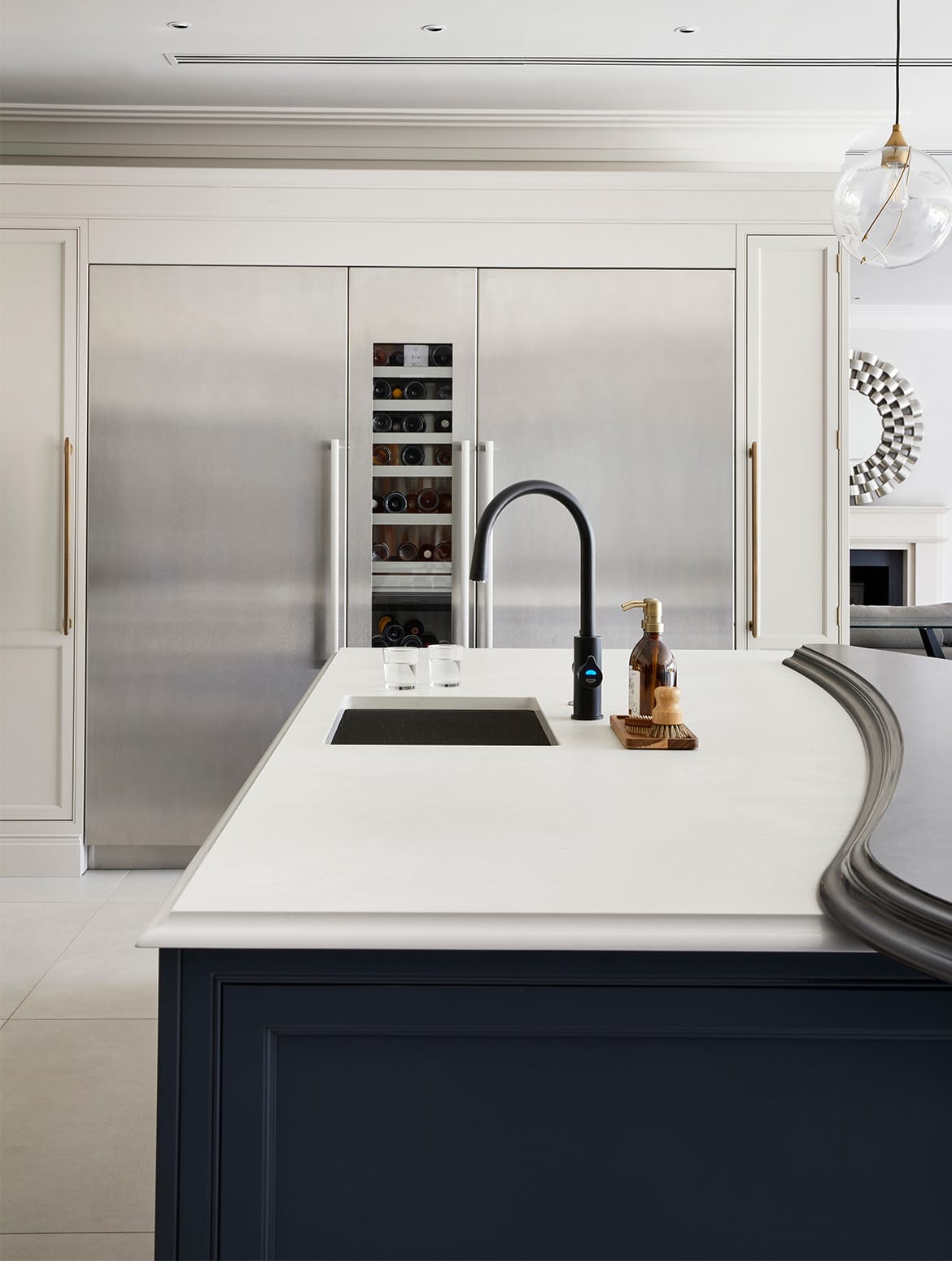 A traditional shaker kitchen project in off-black. A central island with the bespoke cabinetry and a streamlined wood ledge contrasts the white worktop of the remaining island beyond. Against the opposite wall, two Liebherr monoliths are set within French Grey bespoke cabinets, while central to the view is a black Zip tap and accompanying dish soap and scrubbing brush. 