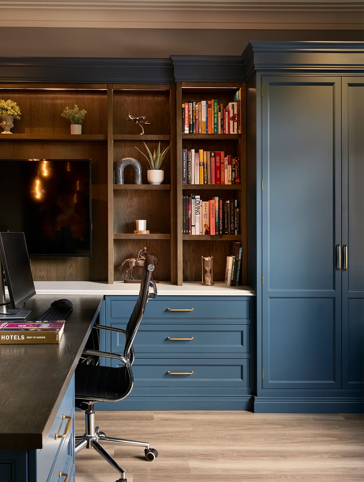 A bespoke home office in off-black. The bespoke cabinetry that runs across the whole left wall houses memorabilia, books, and a mounted television. The TV oversees a desk with space for two people to work opposite each other. The burnished brass handles of the drawers beneath the shelves and the cabinet at the end stand out well.