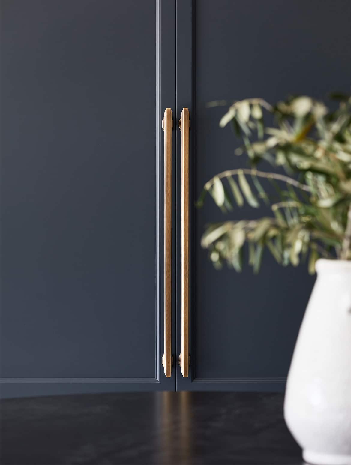 A traditional shaker kitchen project in off-black. A central island with the bespoke cabinetry and a streamlined wood ledge contrasts the white worktop of the remaining island beyond. A ceramic vase overlooks the off-black cabinet doors beyond. 