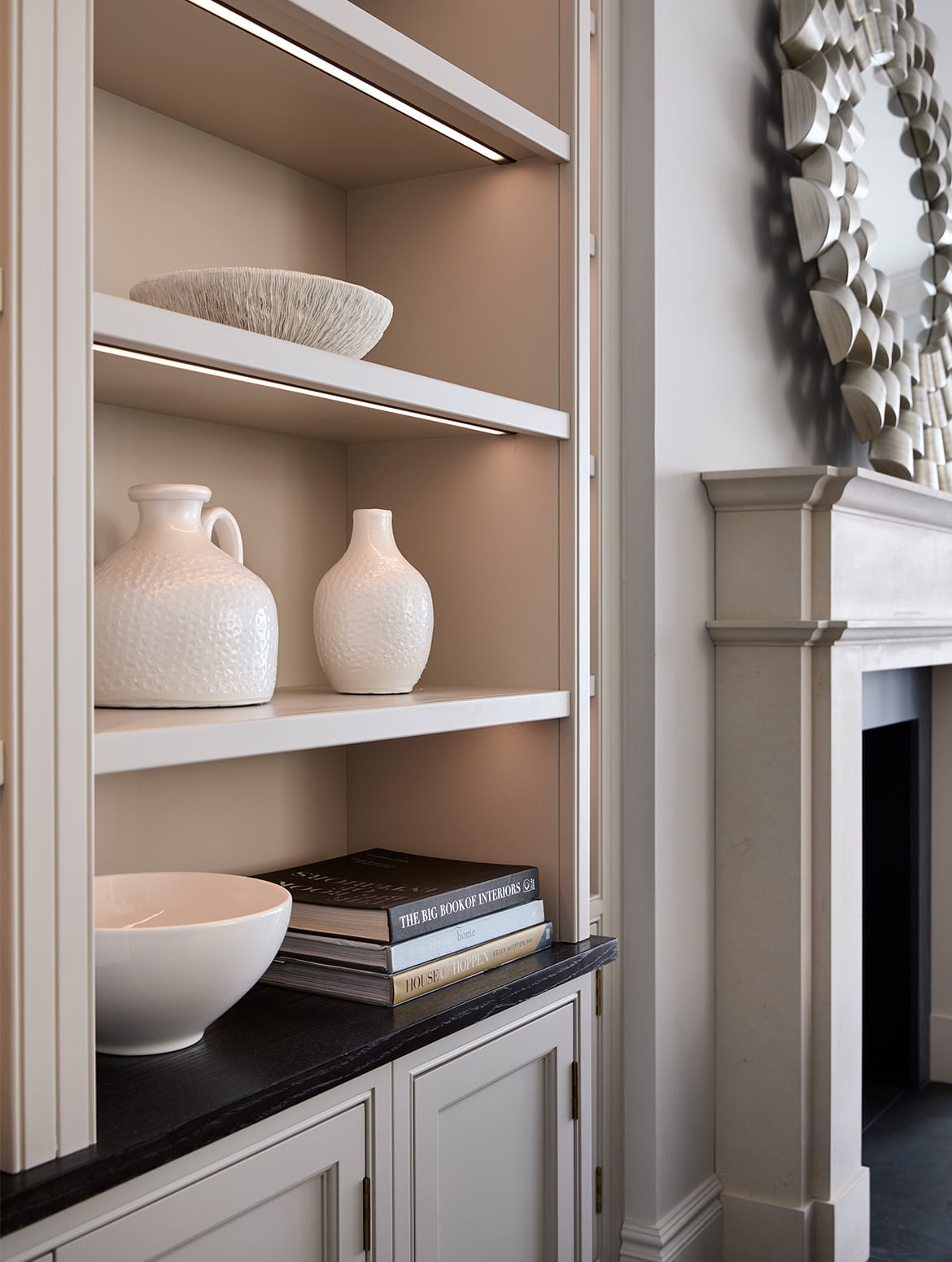 A media wall with bespoke cabinetry in French Grey. A central fireplace with log burner is set within the Edwardian-style mantelpiece beyond bespoke cabinetry set into its left side is visible. The shelves house a ceramic urn, two vases, and a bowl below. Push-to-open cabinetry is just visible below. The concealed lighting in the shelves bathes the items in a warm light. 
