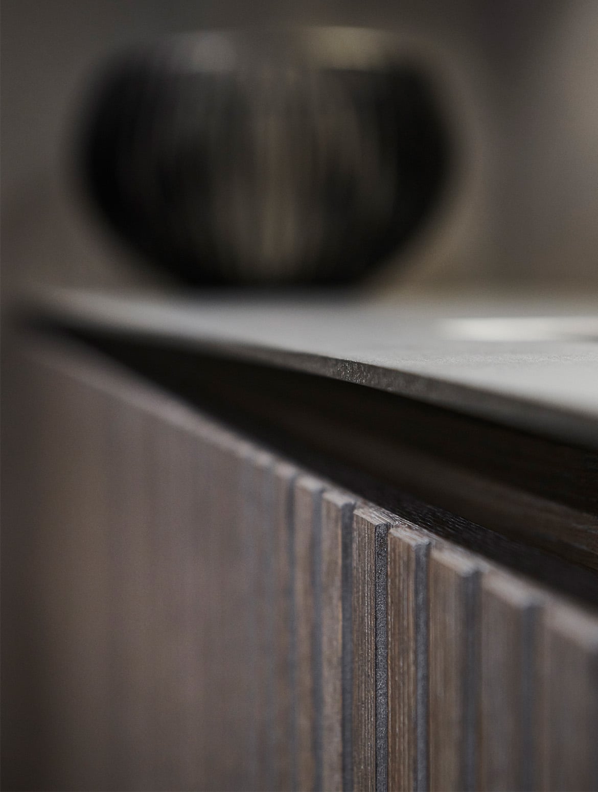 A modern kitchen in smoked wood finish. A close in view of the flush lined bespoke cabinetry with a smoked wood finish. Though blurry, a brown ceramic urn stands on the ledge. 